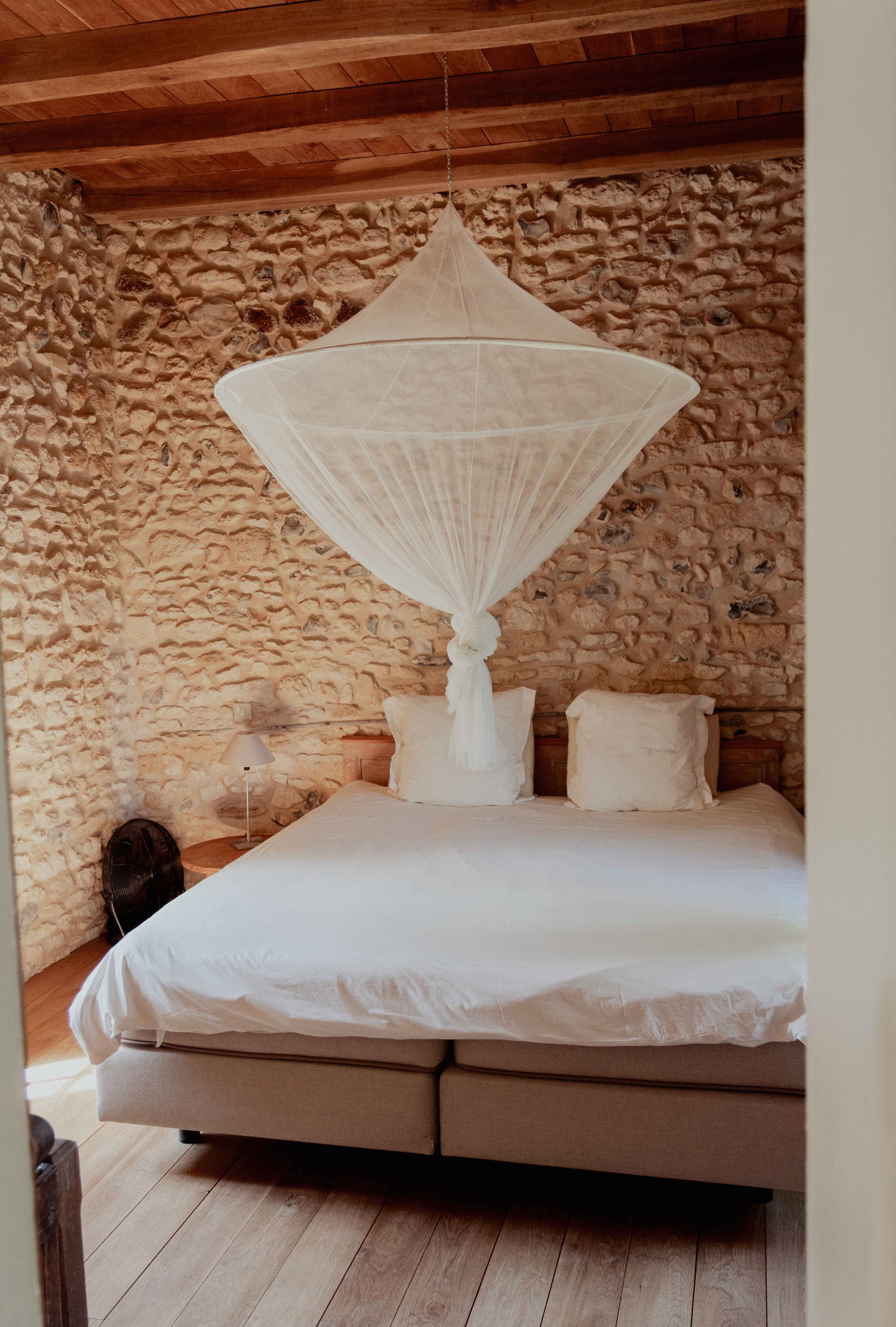 A bedroom with a bed, stone wall, wooden ceiling, and a white fabric lantern hanging above the bed.