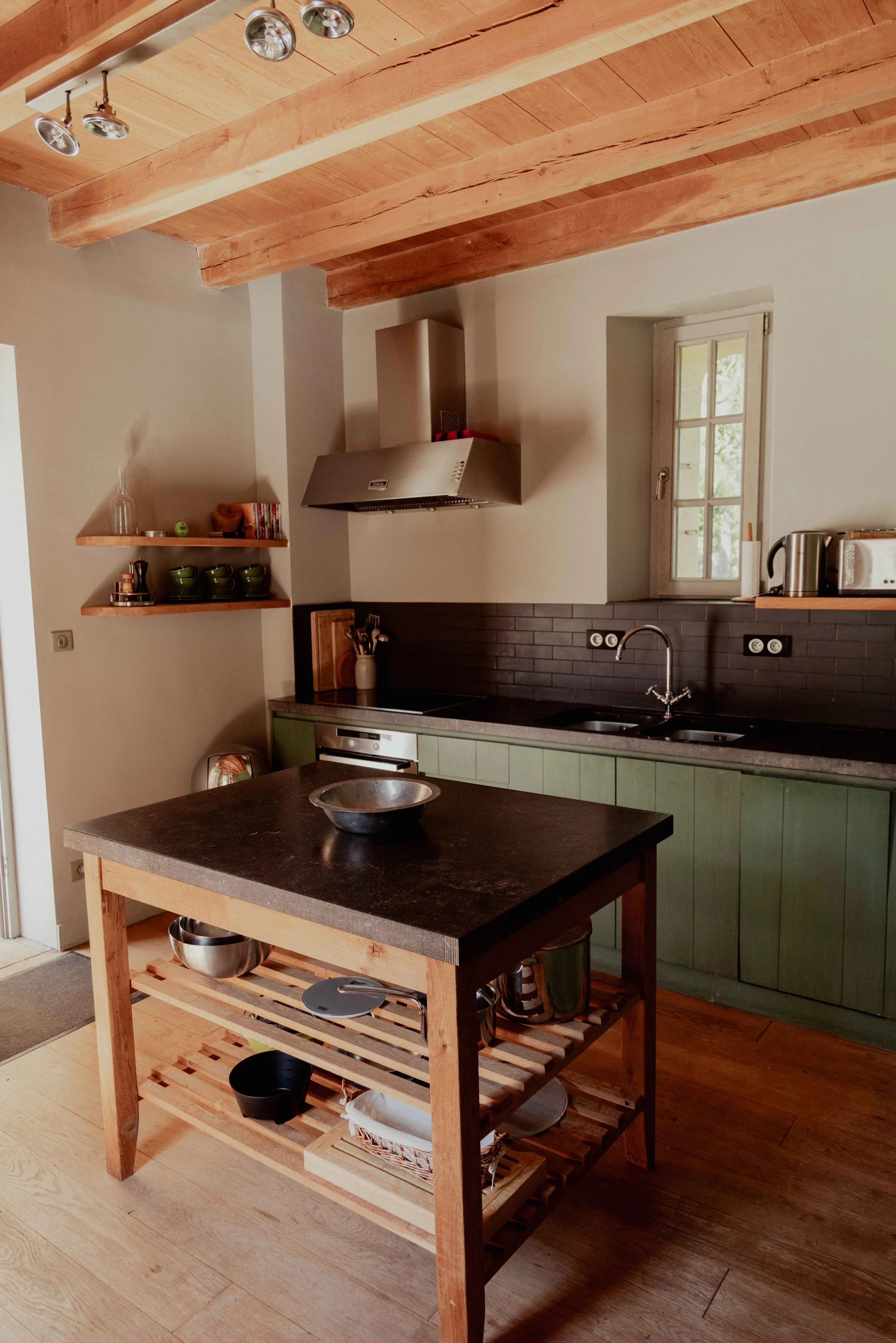 Een keuken met een houten plafond en een keukentafel, groene keukenkastjes, een ramen, een schouw, en open planken met servies en keukenbenodigdheden.