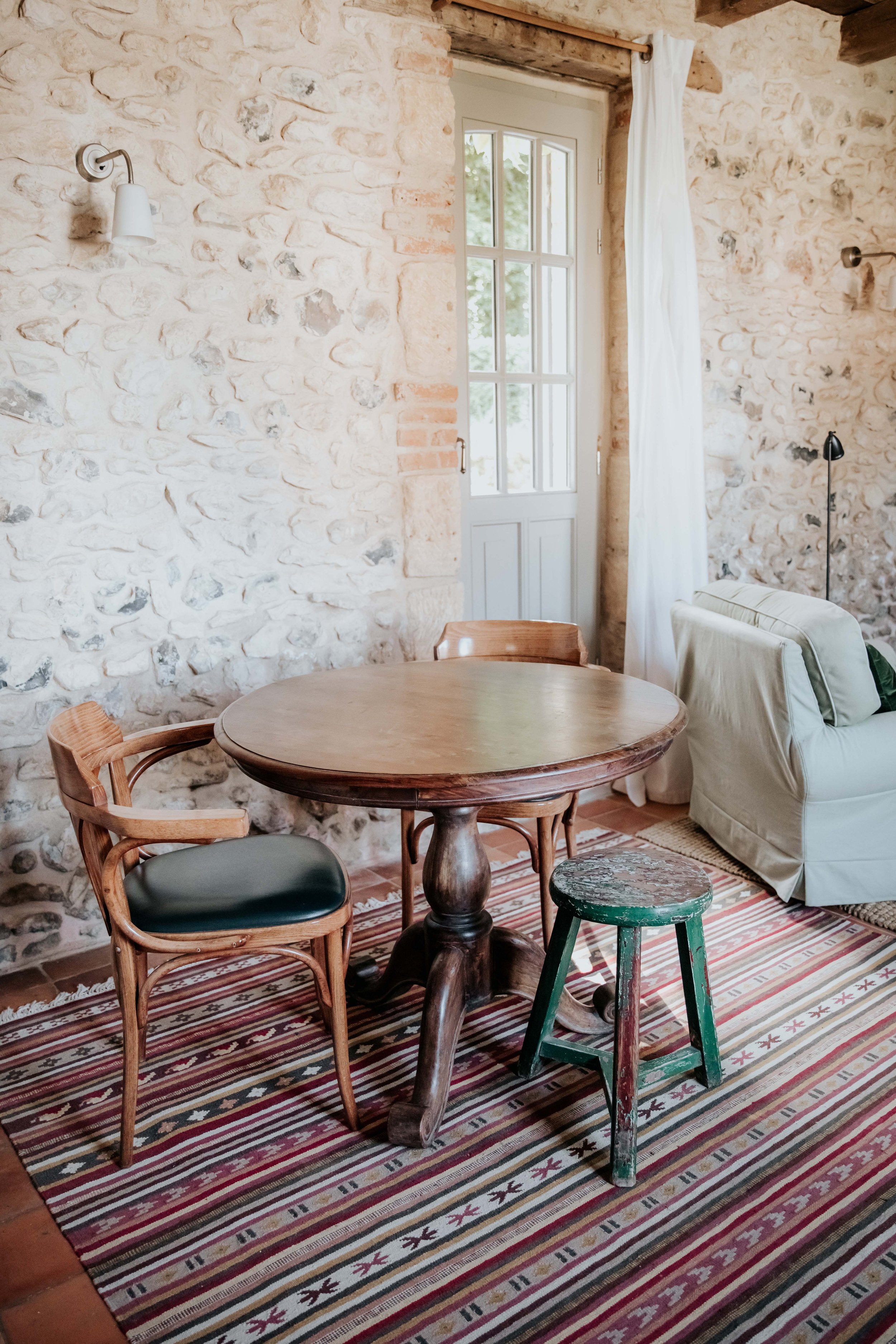Interior of a rustic room with a stone wall, wooden table, and mixed seating including a wooden chair with a blue cushion, a green painted stool, and a white sofa. A window with white curtains lets in natural light.