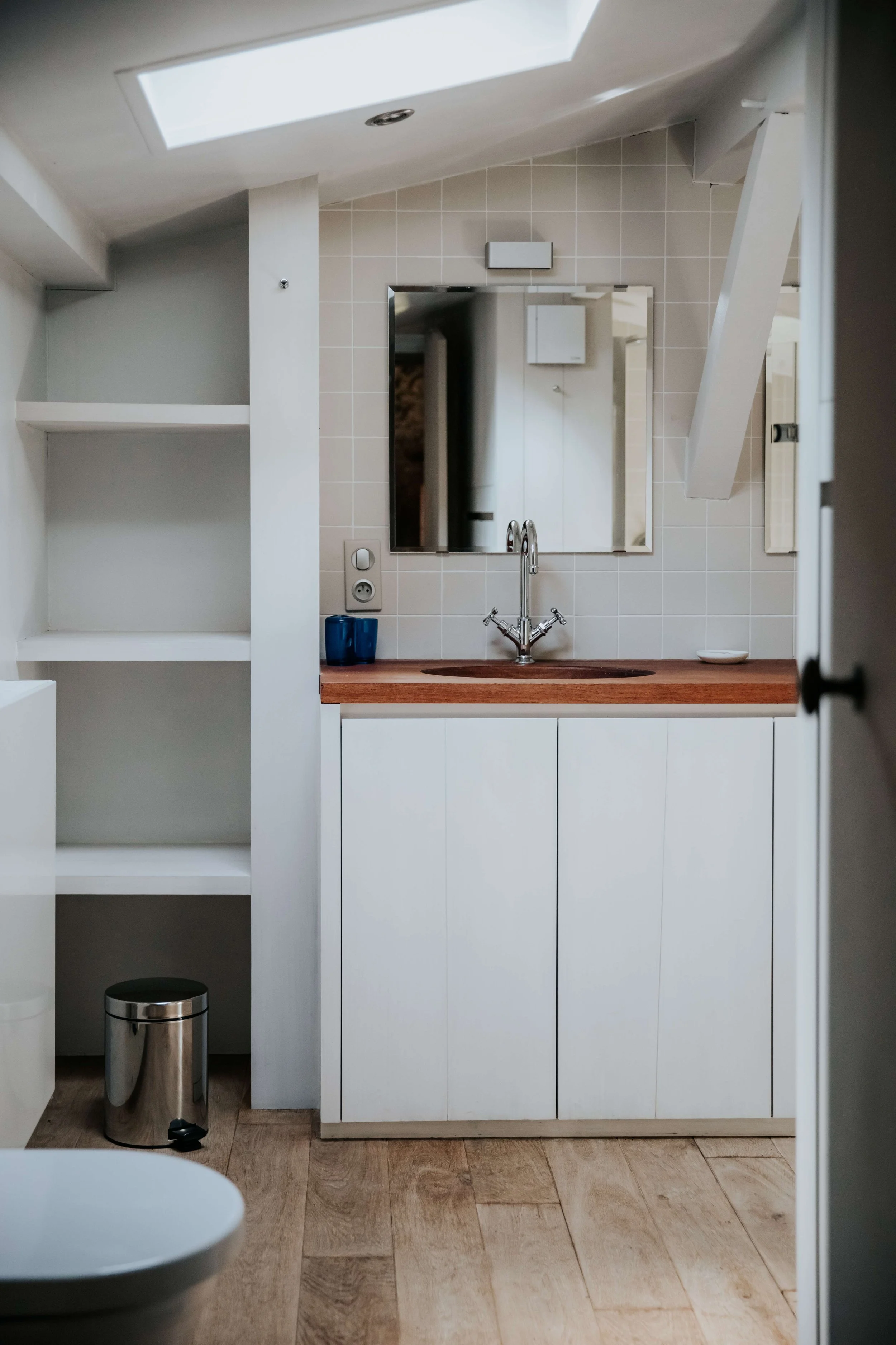 Small modern bathroom with a skylight, a white vanity cabinet with a wooden countertop, a small sink with a chrome faucet, a mirror, a blue cup, a white soap dish, beige tiled wall, open white shelves to the left, and a small trash can on wooden floor.