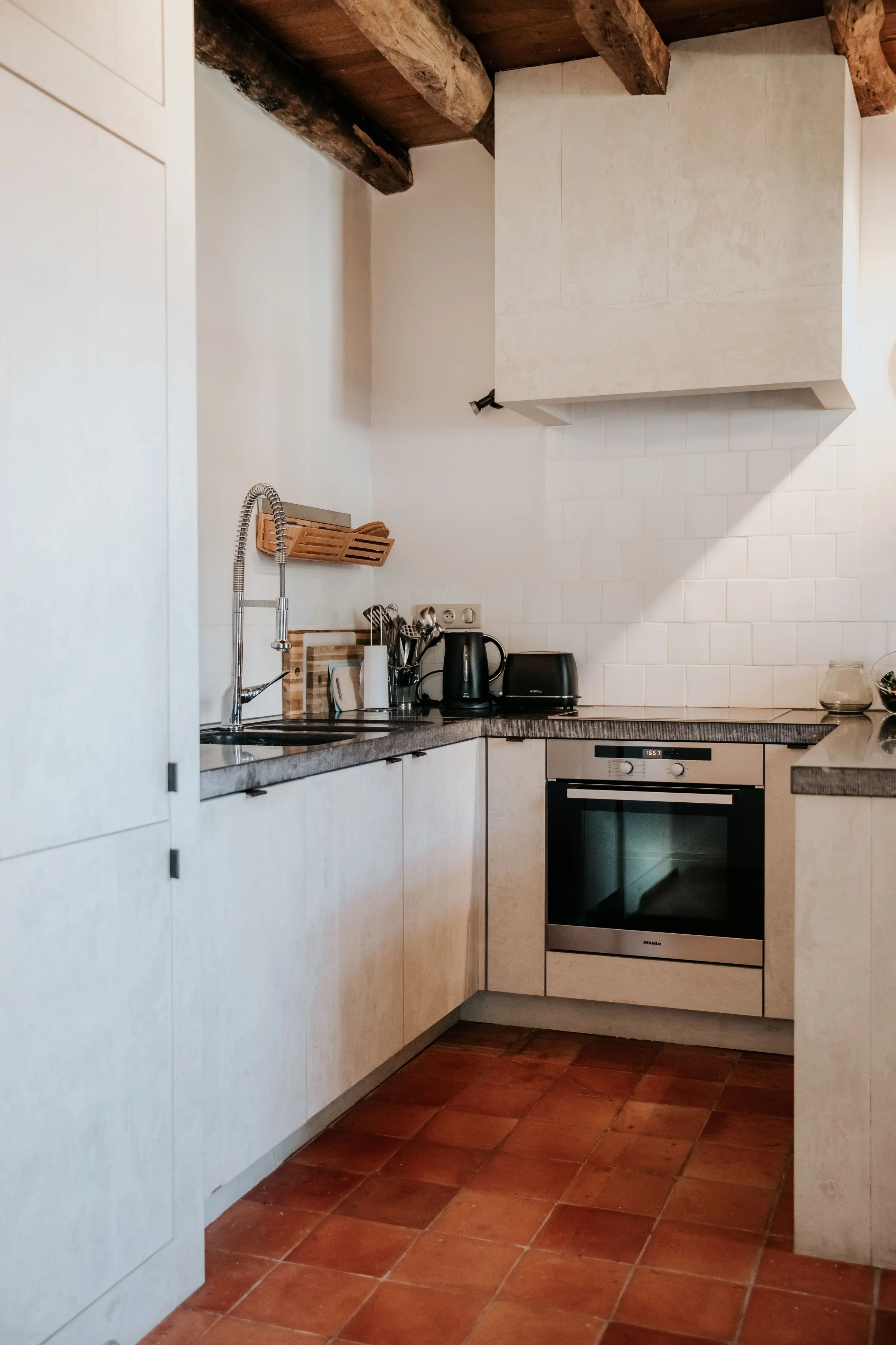 A modern kitchen with white cabinets, a dark countertop, a stainless steel oven, and various kitchen appliances including a kettle, toaster, and utensils. Wooden beams are visible on the ceiling and the floor is made of terracotta tiles.