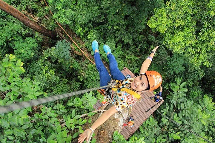Een meisje hangt aan een touwbrug hoog in de bomen, omringd door een dicht groen bladerdak, lachend naar de camera, met een klimhelm en klimuitrusting.