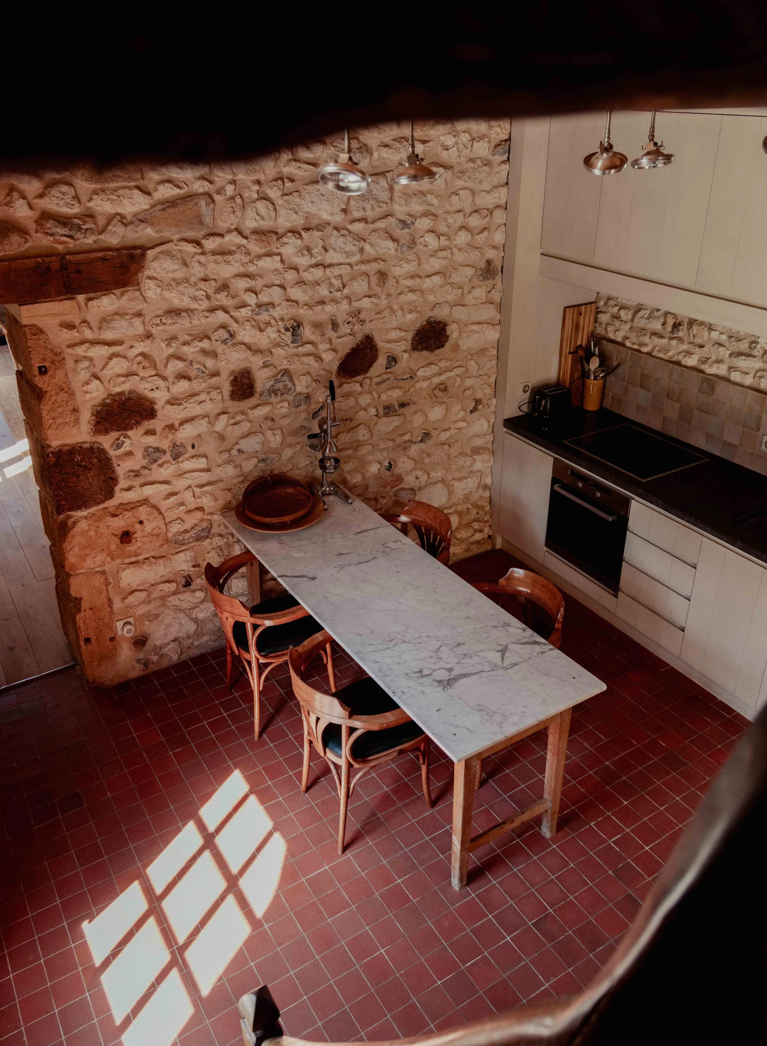 Uitzicht op een keuken met een lange marmeren tafel en vier houten stoelen, een bakstenen muur en moderne plafondlampen.