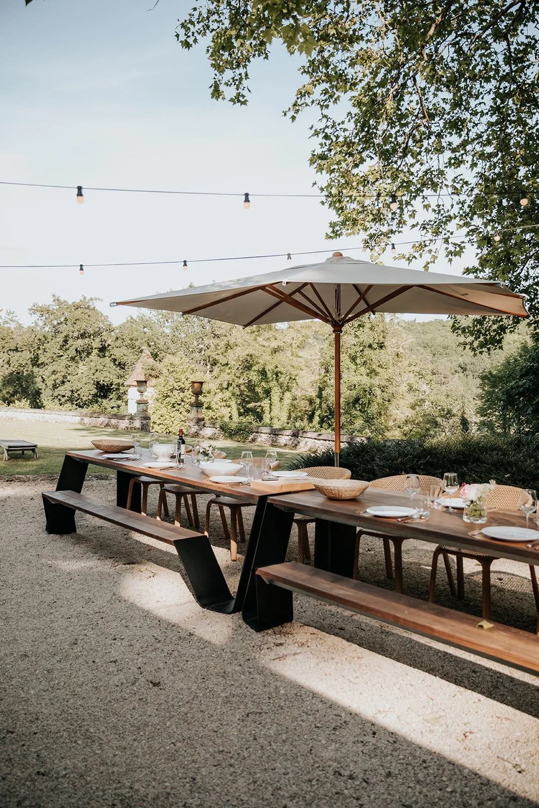 Outdoor dining table set up with plates, glasses, and bowls, shaded by a large umbrella, outdoors with trees and string lights overhead.