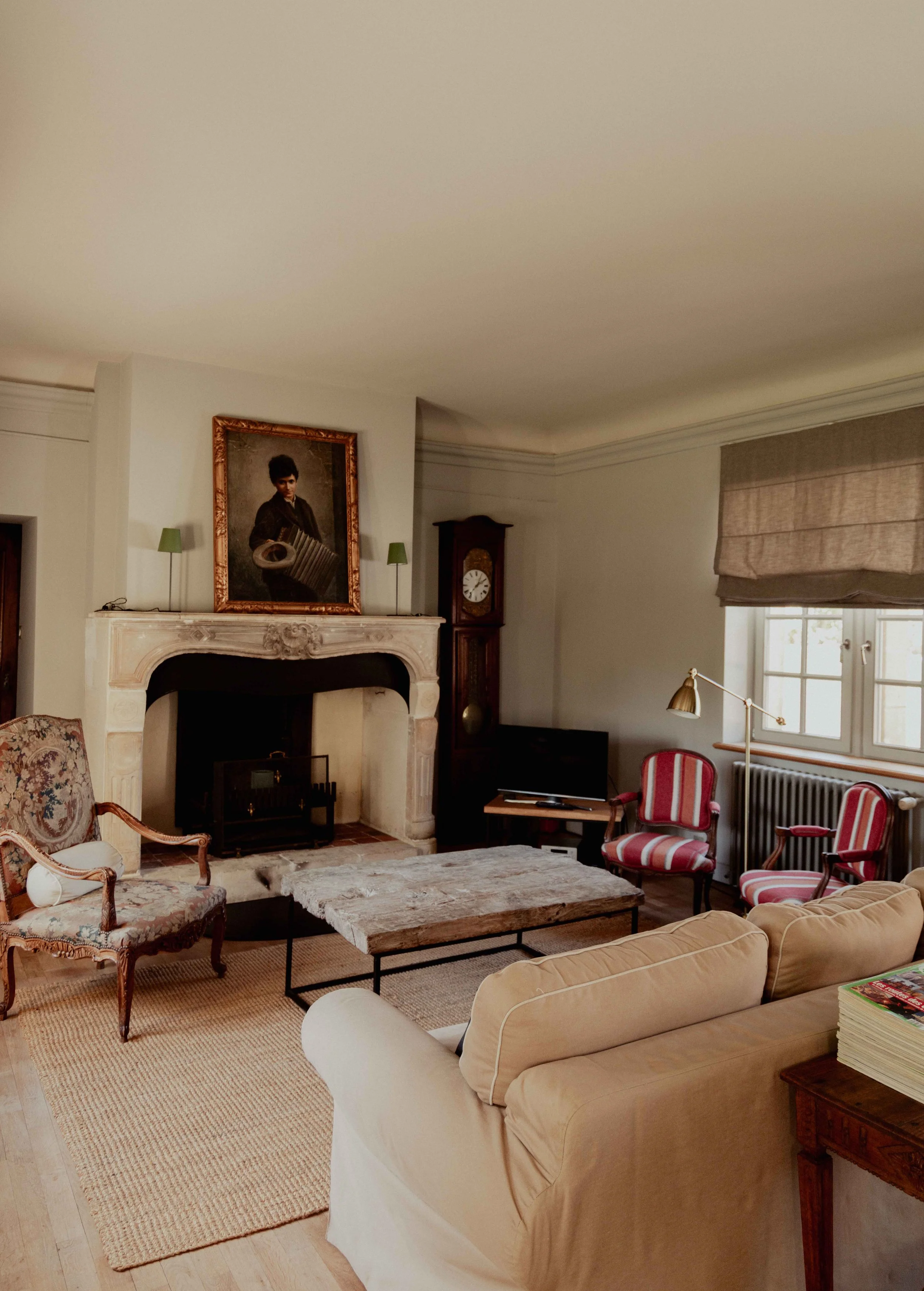 Gezellig woonkamer met antique meubels, een open haard met een foto boven, een grote beige bank, en een televisietoestel. Rood gestreepte stoelen, een vloer met een paar tapijten en een raam met een beige raamkleed.