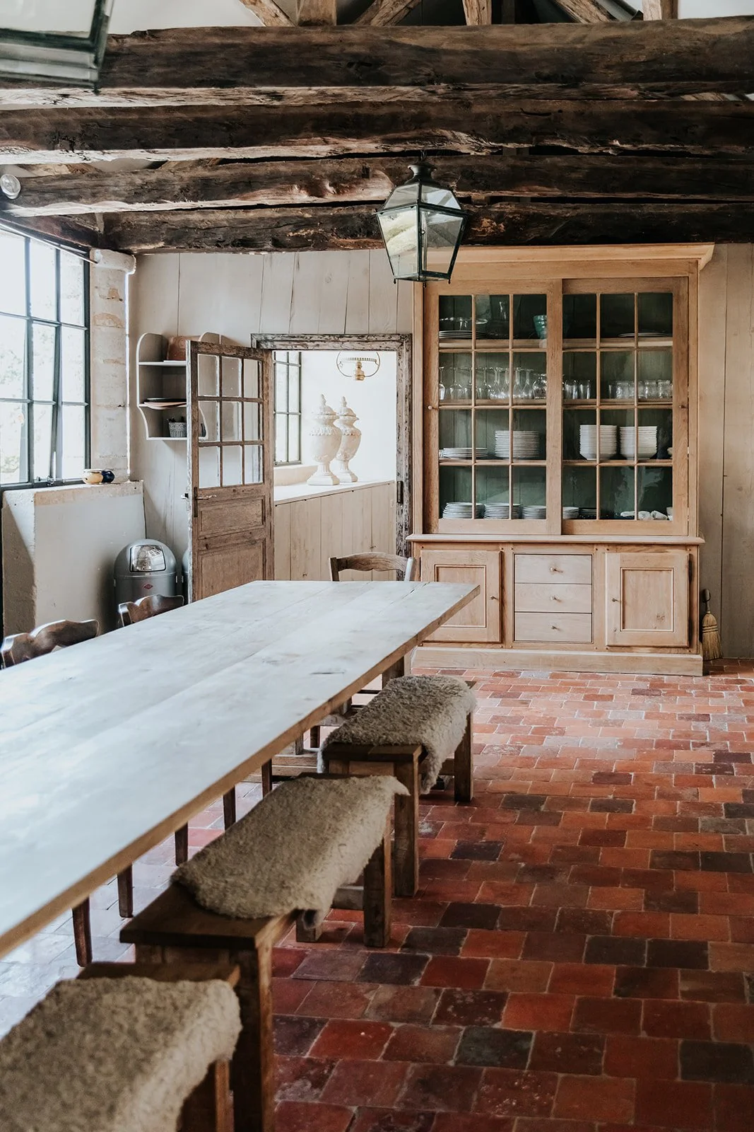 A rustic dining room with a long wooden table, four chairs with beige furry cushions, a large glass-fronted wooden cabinet filled with dishes, brick tile flooring, and exposed wooden ceiling beams. There are large windows on the left letting in natural light.