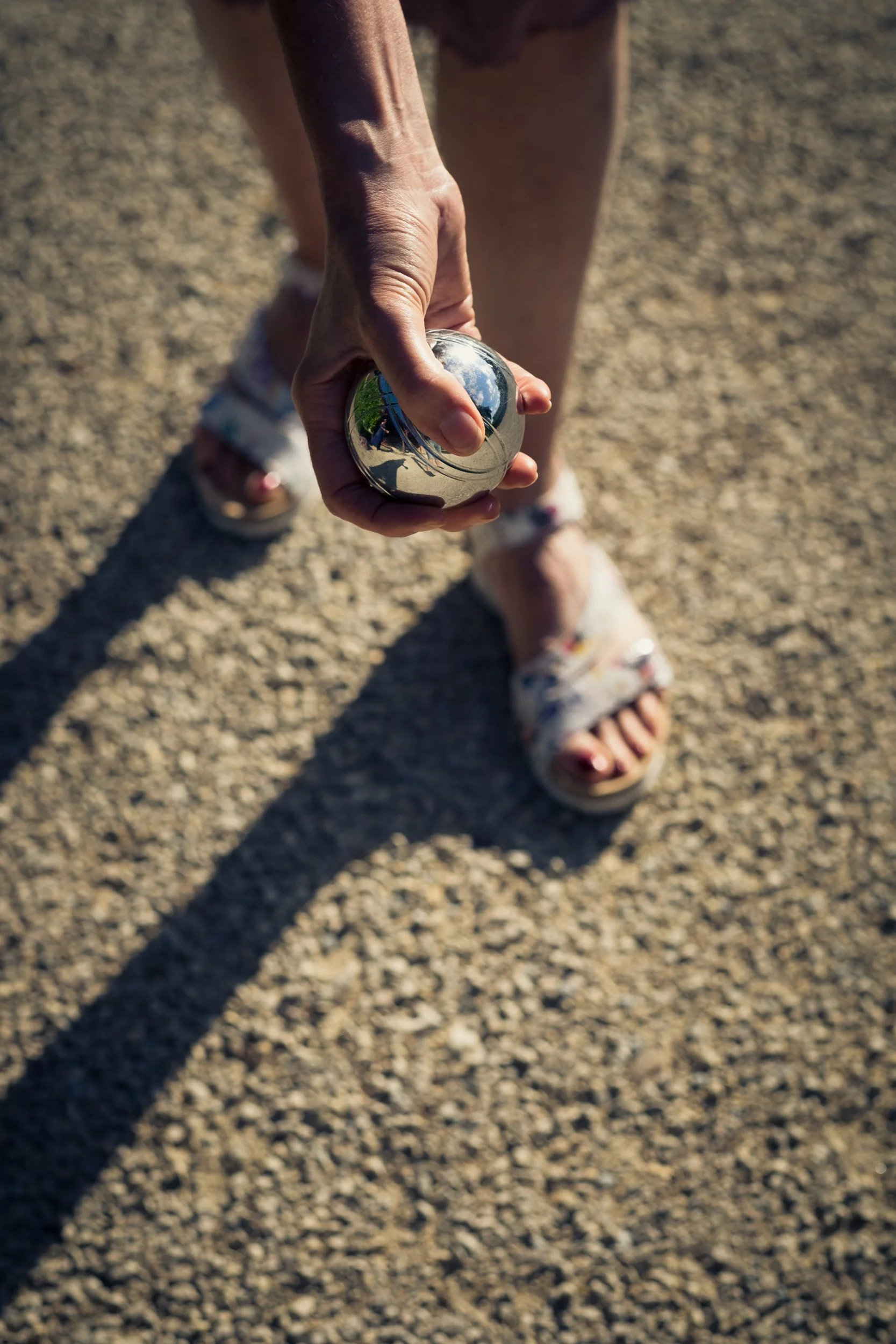 A person holding a shiny metal ball, with the reflection visible, while standing on a gravel surface, wearing floral open-toe sandals.