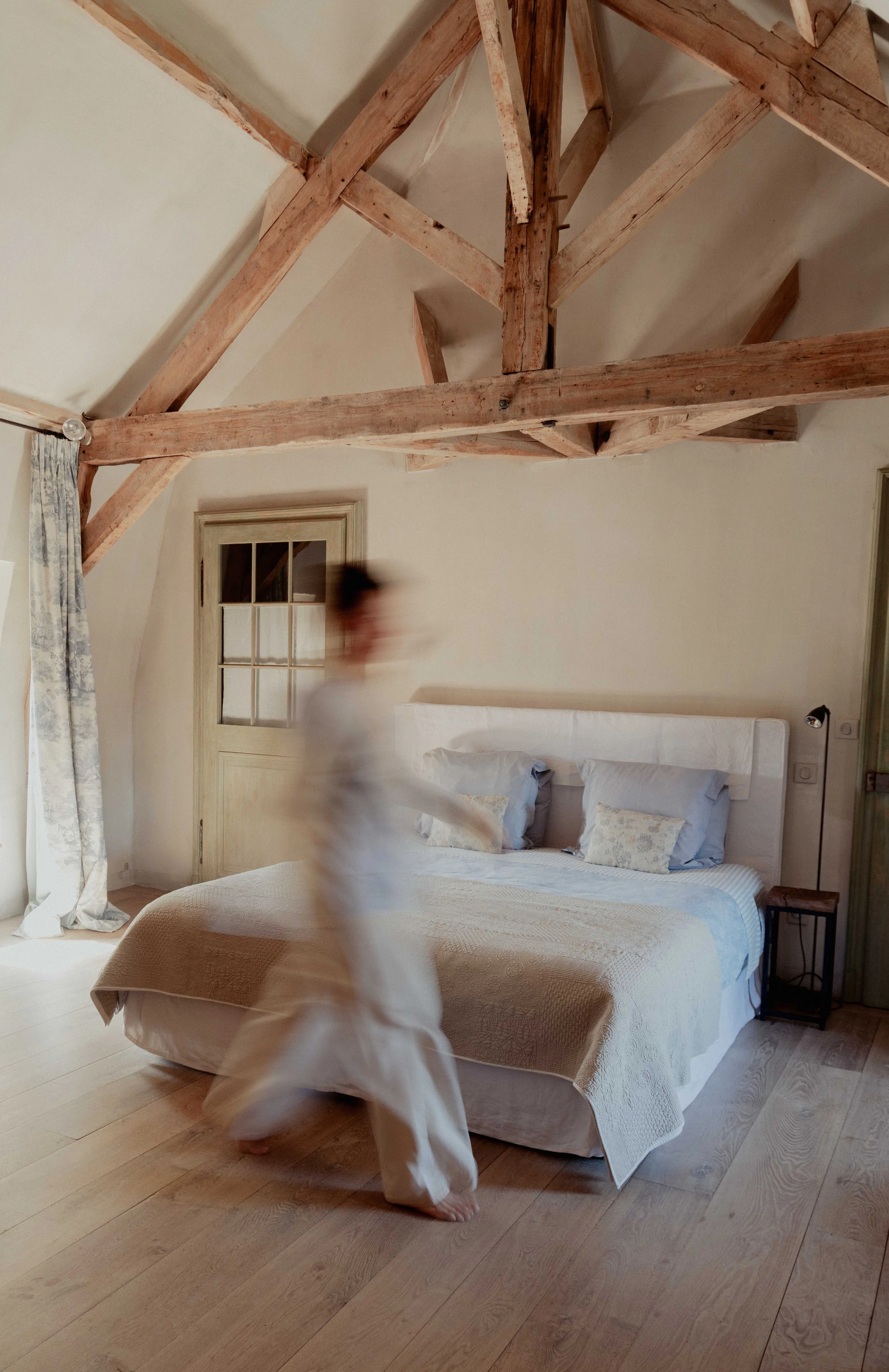 Een persoon die snel door een slaapkamer met een hoog plafond loopt, met zichtbare houten balken en een bed in het midden.