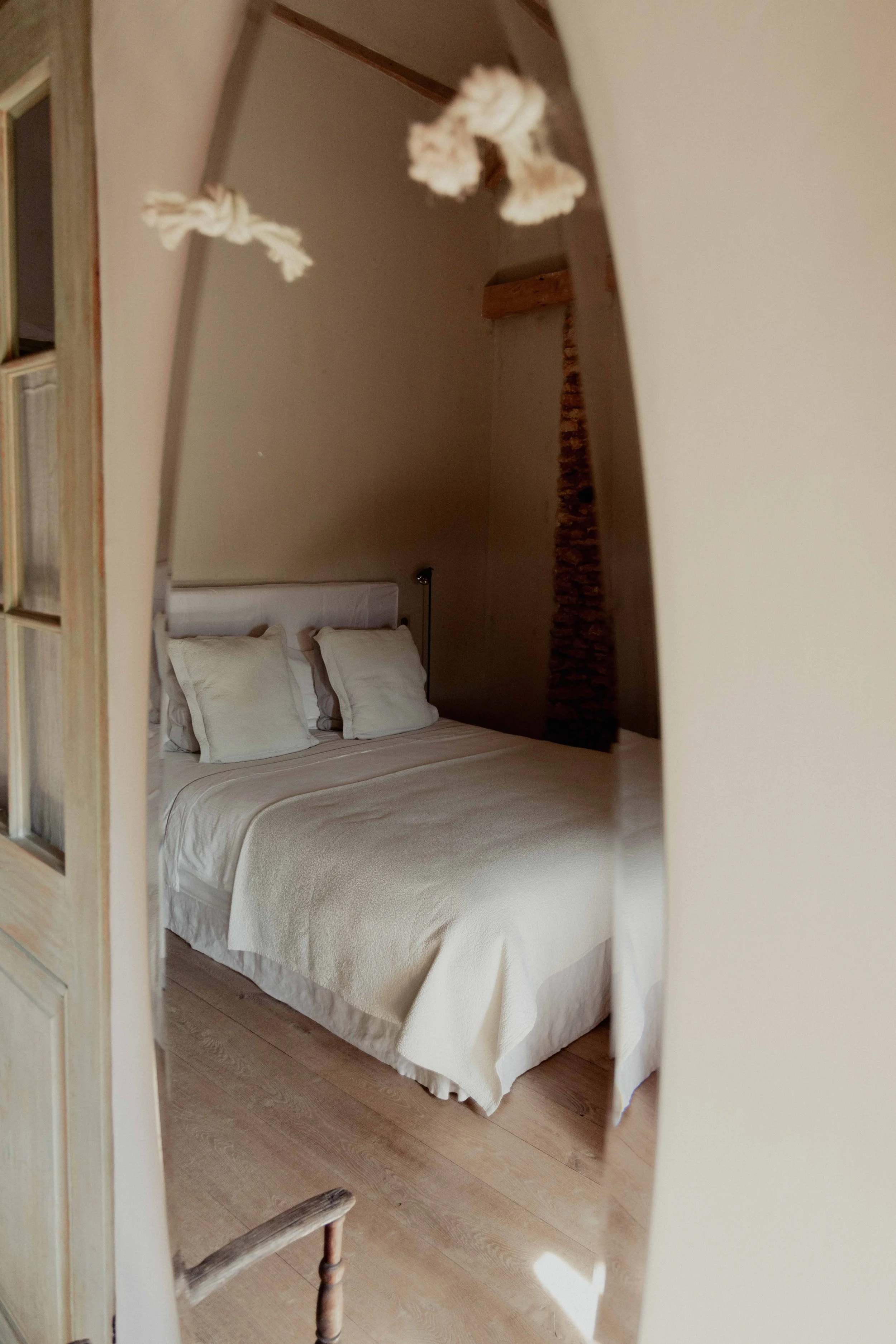 Een wit bed met twee witte kussens op een houten vloer, gezien door een scheef spiegel in een kamer met een schuine plafond en een deels open deur.