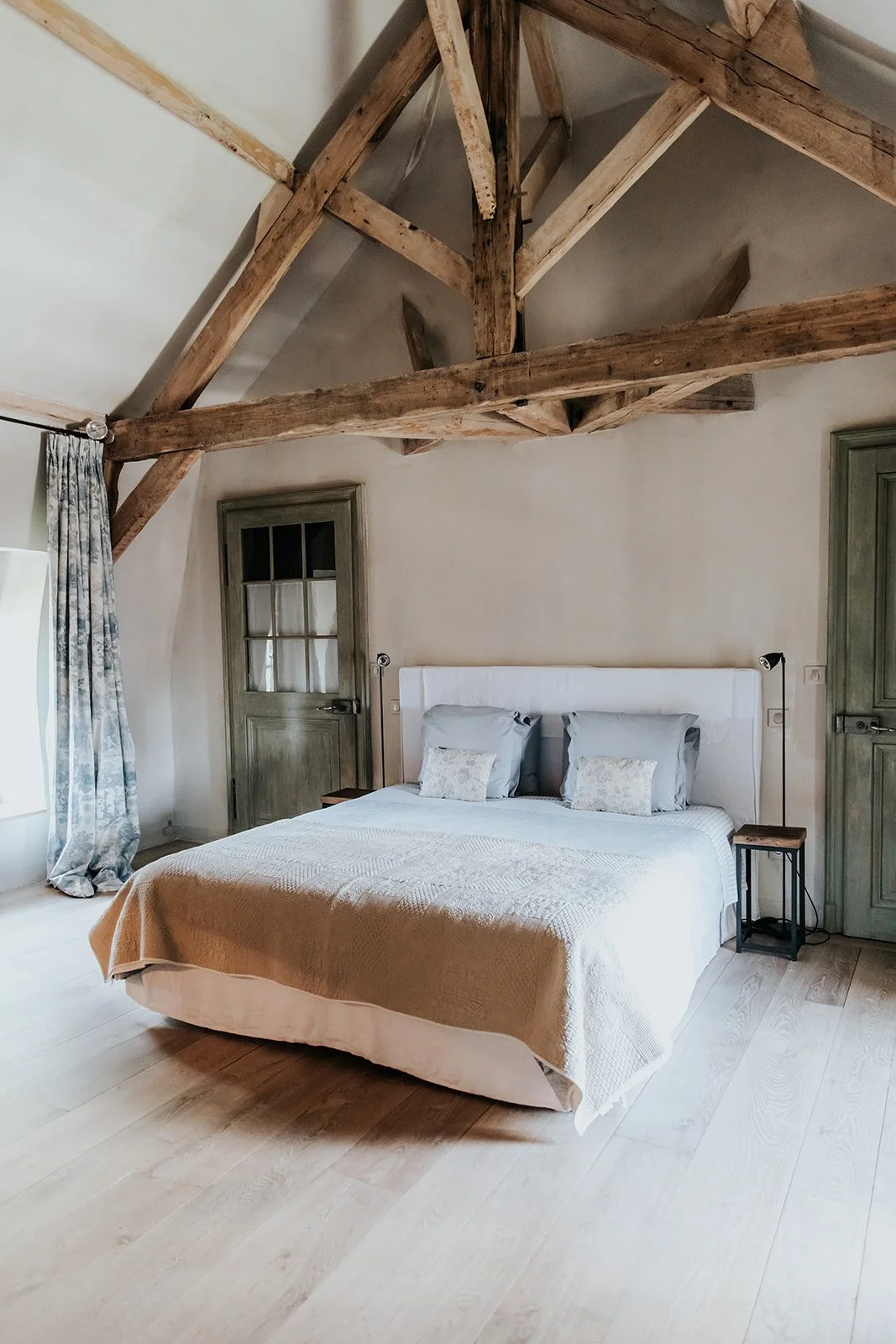 A bedroom with a vaulted ceiling featuring exposed wooden beams, a bed with a beige quilt and three pillows, wooden nightstands, a door with a glass panel, and a window with patterned curtains.