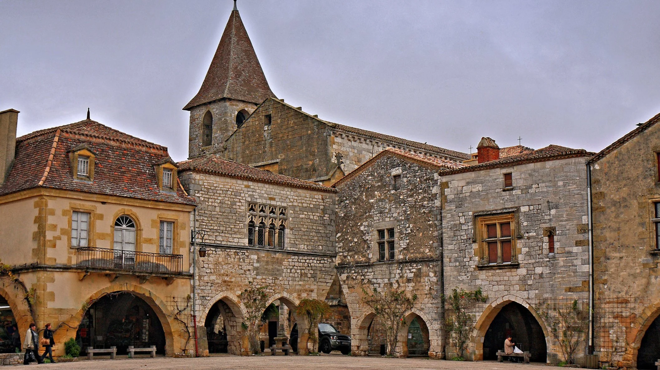 Historic European town square featuring stone and stucco buildings with arched arcades on the ground level, a church with a tall spire, and a cloudy sky overhead, with pedestrians and parked cars.
