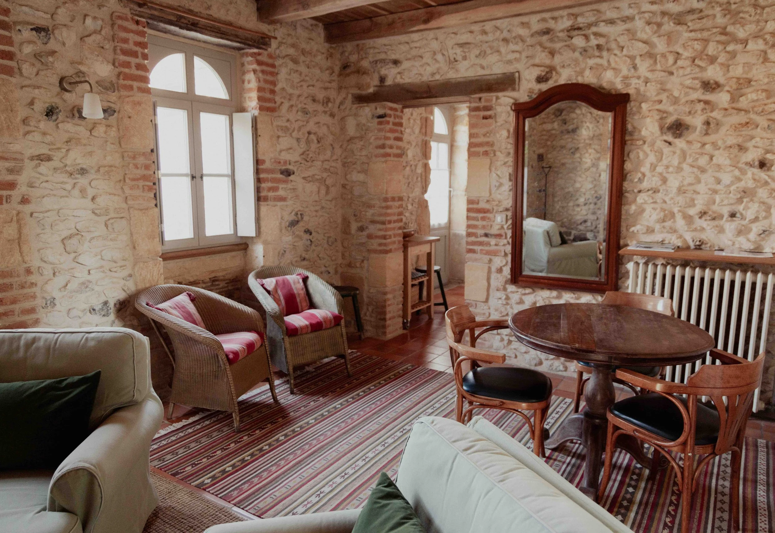 Een woonkamer met stenen muren, houten balken en grote ramen met witte kozijnen, ingericht met beige en rood gestreepte banken, rieten stoelen met kussens, een ronde houten tafel, een grote spiegel in een houten frame, en een radiator.