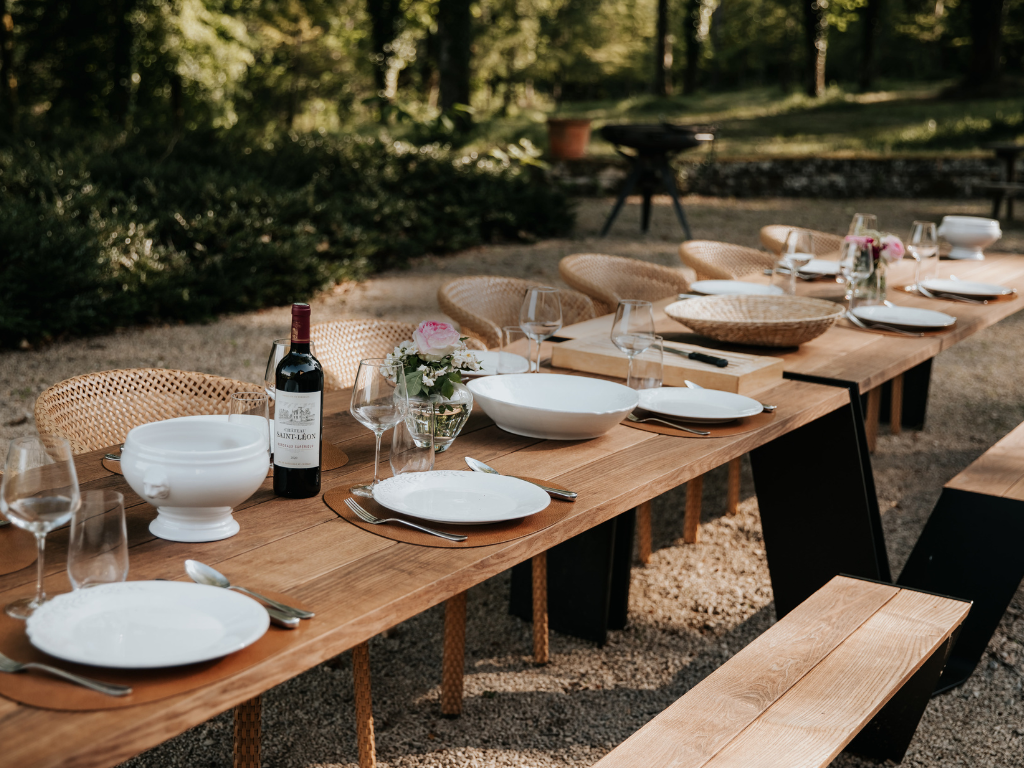 Een lange houten tafel met wit servies, glazen, bestek, wijn en bloemstuk, opgesteld buiten in een tuin of park.