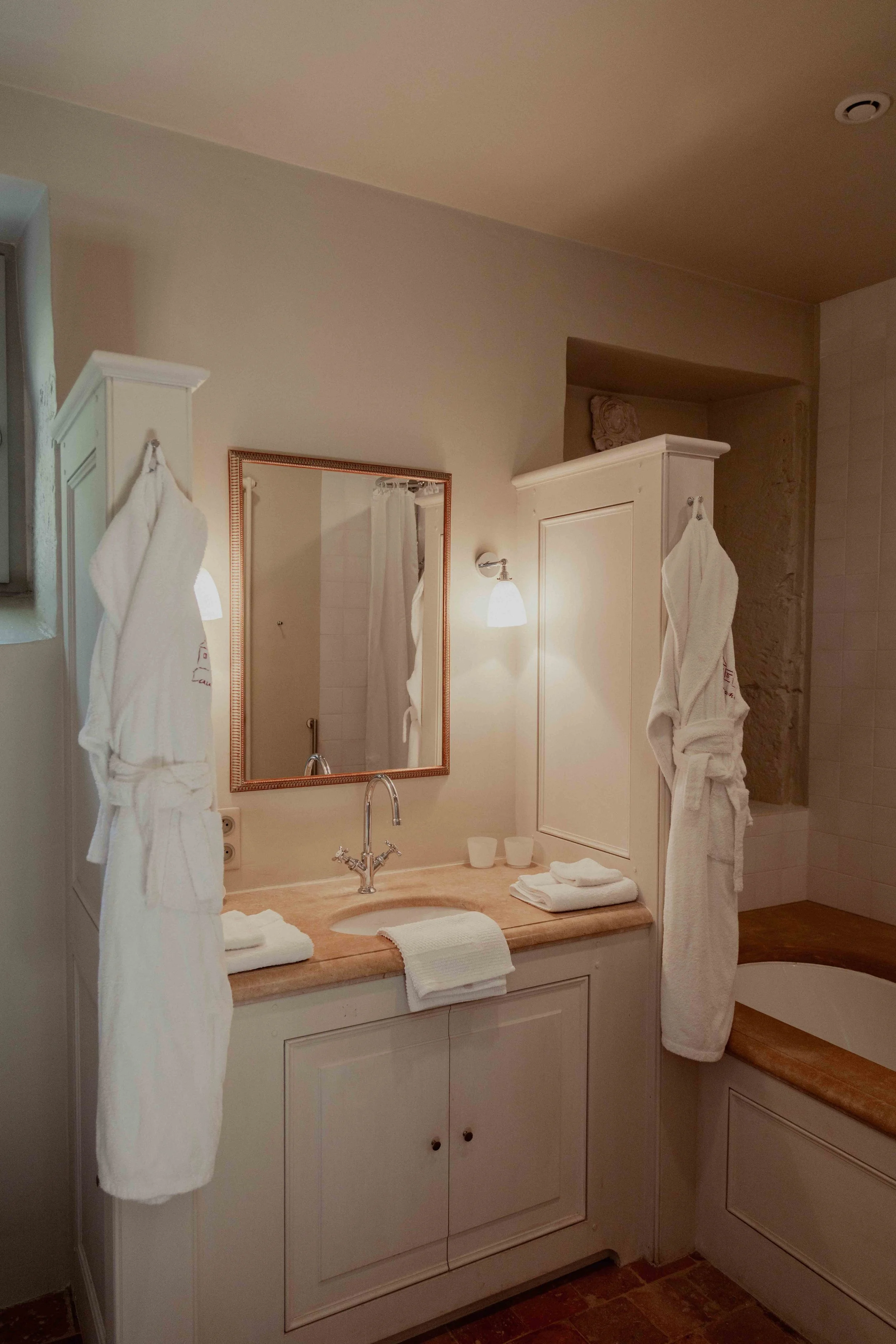 A bathroom with a cream-colored vanity, mirror, two wall-mounted lamps, and two white bathrobes hanging on cabinets. There are folded towels on the countertop, and a bathtub partially visible on the right side.