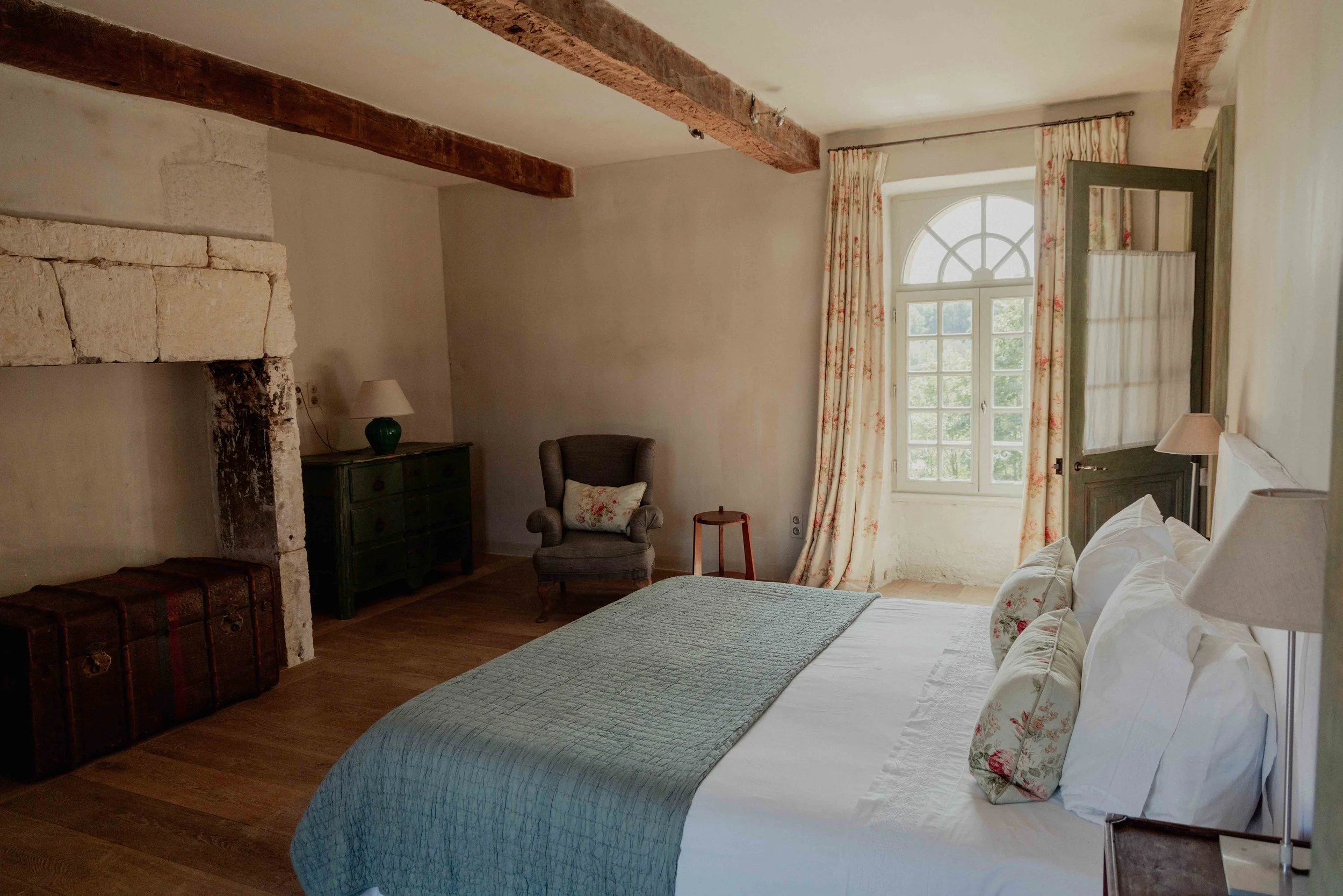 A cozy bedroom with a large bed, white linens, floral pillows, and a green quilt, near a window with floral curtains, a wooden floor, a vintage armchair, a small table, a chest, a dark green dresser with a lamp, and exposed wooden ceiling beams.