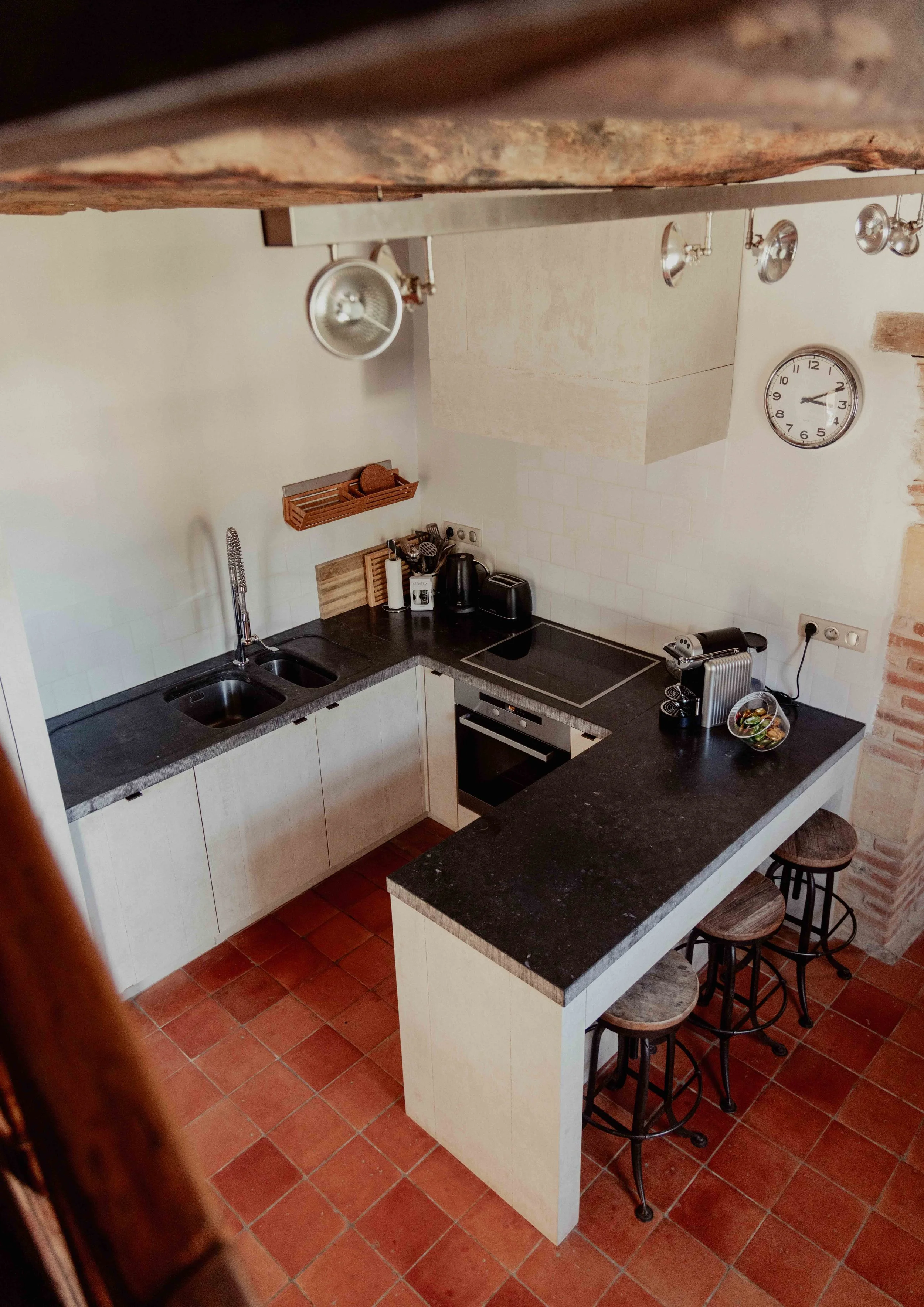 Kleine keuken met zwart werkblad, wit keukenkastjes, dubbele spoelbak, kookplaat, oven, koffiezetapparaat, broodrooster, keurig opgeruimd, klok aan de muur, barstoelen aan eiland, terracotta vloer, lichte muur met bakstenen muur bij de bar.