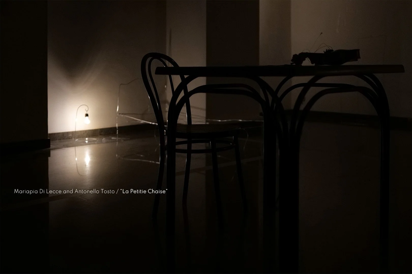 A dimly lit room with a small lamp on the floor casting a soft glow, nearby are dark metal chairs around a table, and objects on the table including a remote control and papers.