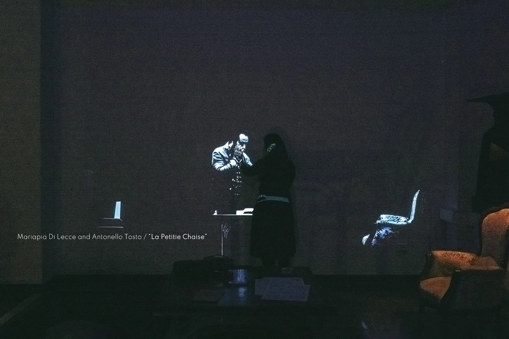 A woman standing in front of a large screen, interacting with a projected image of two men, with a chair and another chair with a floral pattern to the right, and a small table in the foreground.