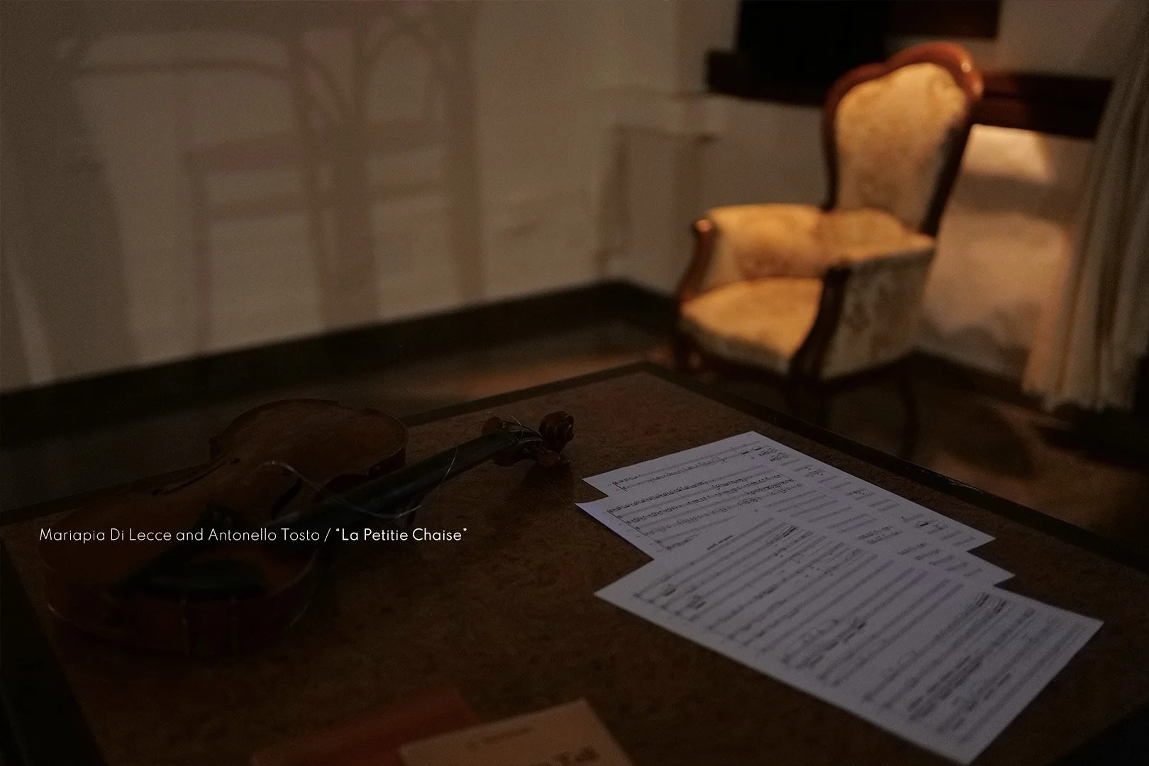 A violin resting on a table with sheet music, with an upholstered armchair in the background. The scene has low lighting.