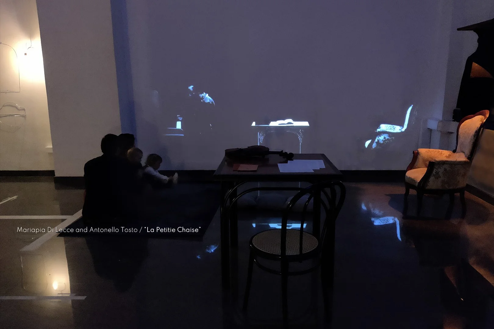 People sitting on the floor watching a movie projection on a wall in a dimly lit room, with vintage chairs and a table visible.
