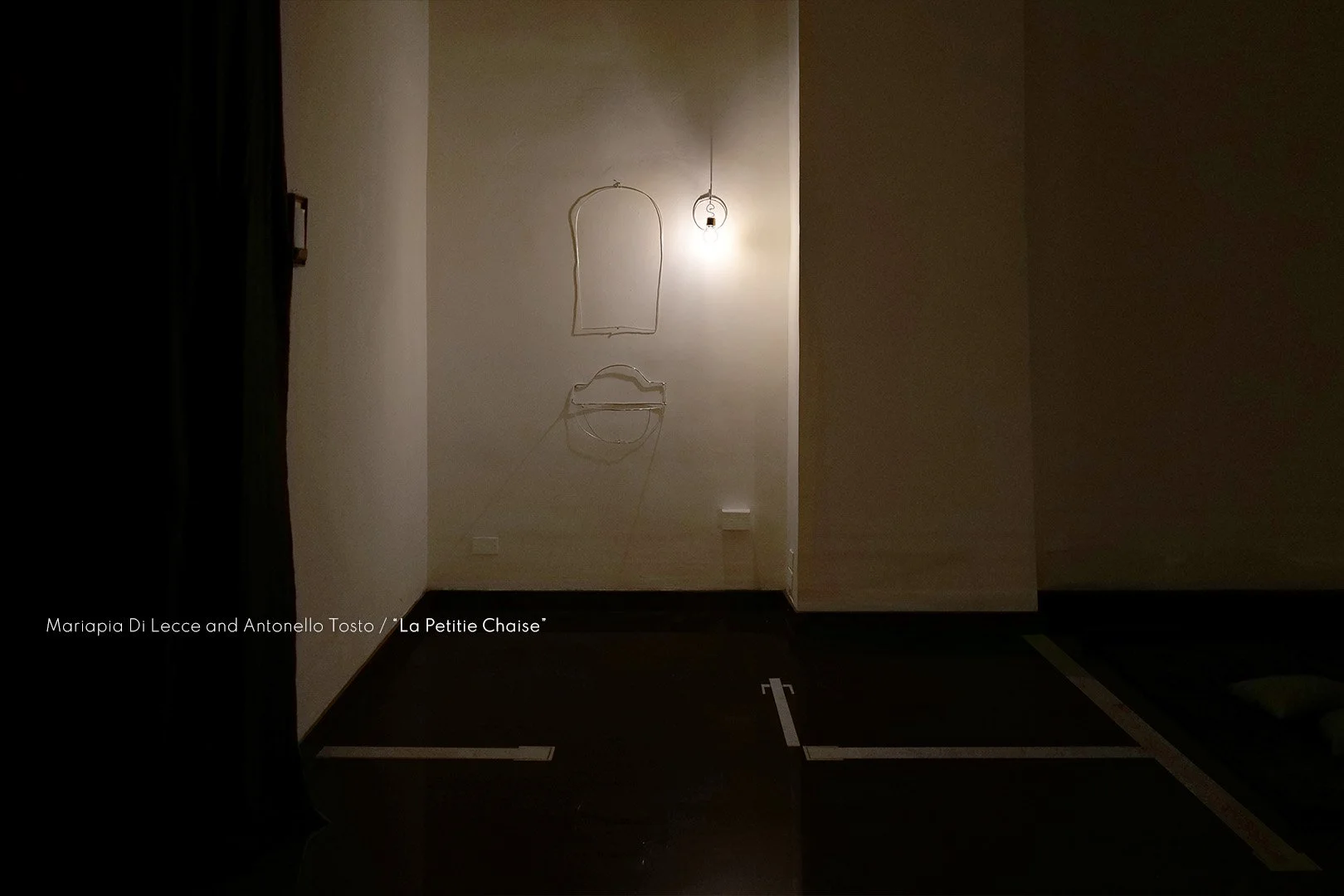 Installation artwork on a wall with a light bulb, consisting of wire outlines of a piece of bread and a small shelf below it.