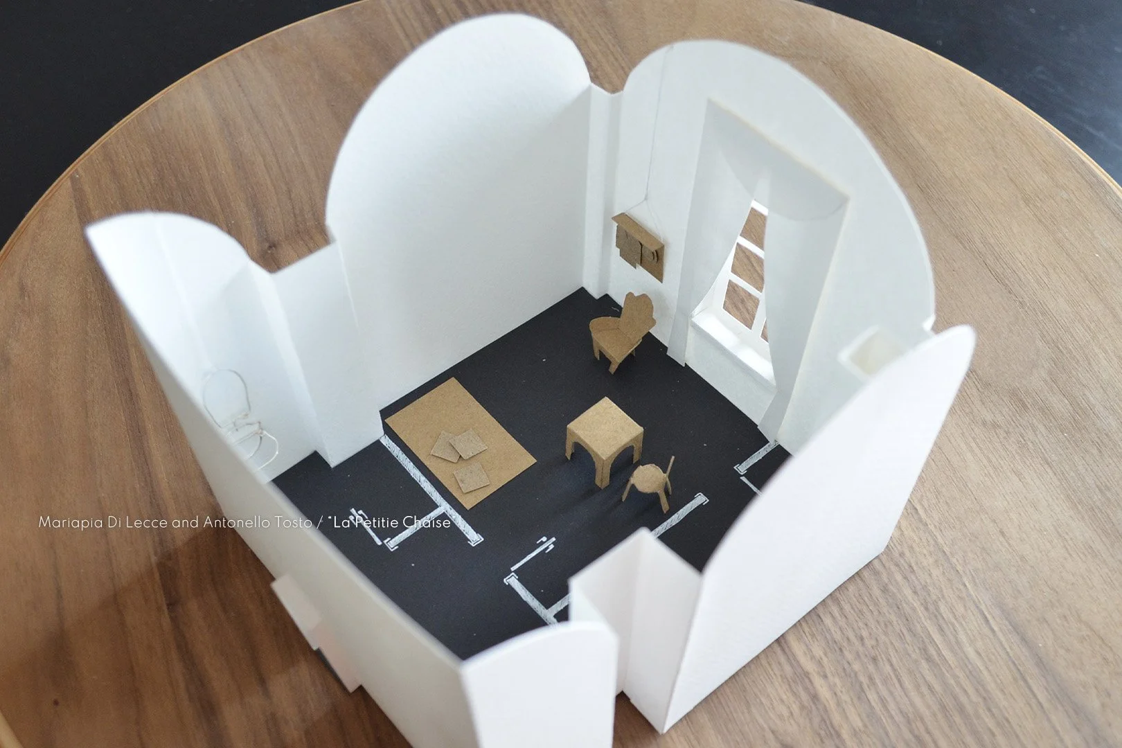 A miniature dollhouse room with white walls, black flooring, and small furniture pieces including a table, chairs, and a little shelf, viewed from above.