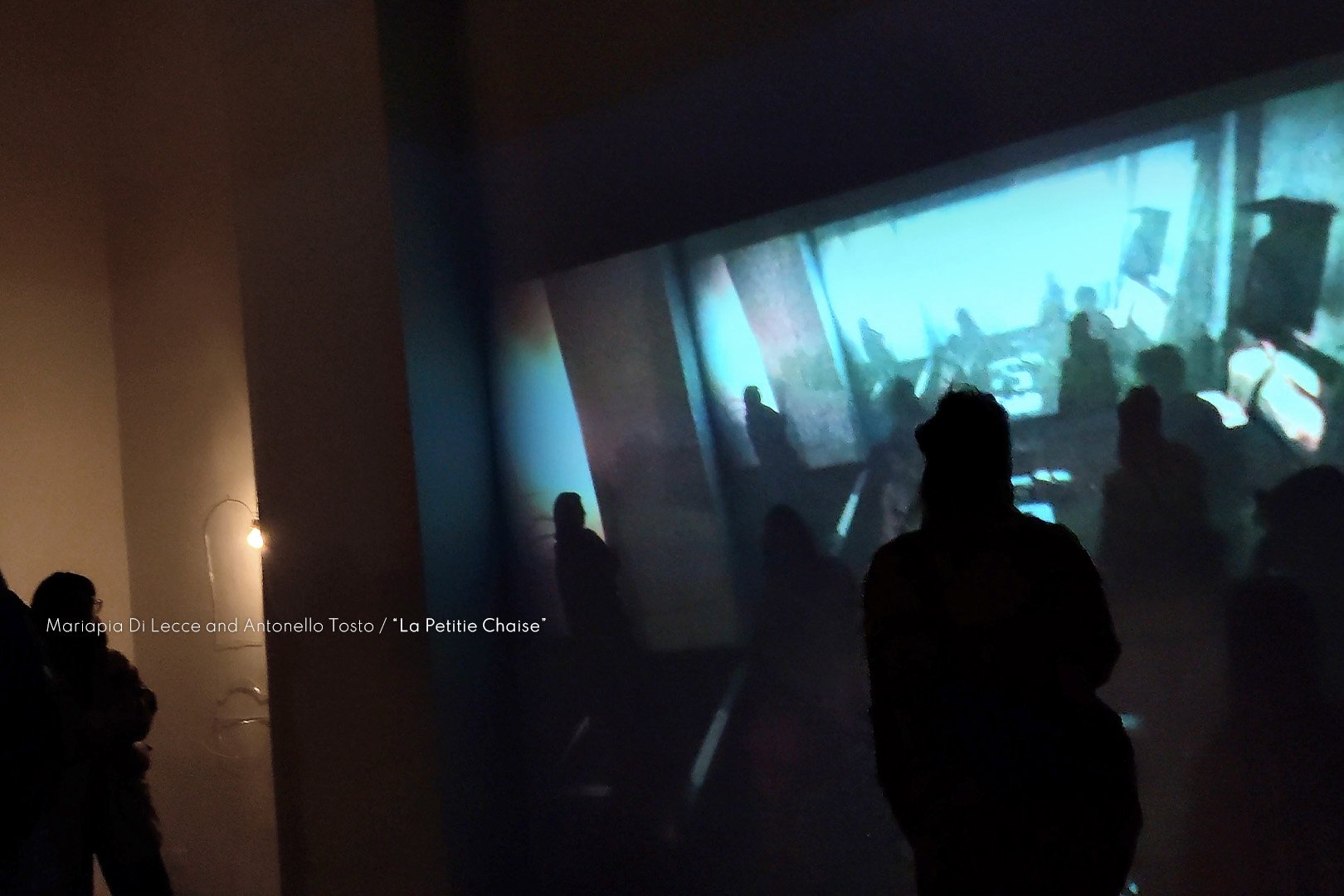 Visitors observing an art installation and video projection in a gallery space, with silhouettes against a large screen displaying an underwater scene.