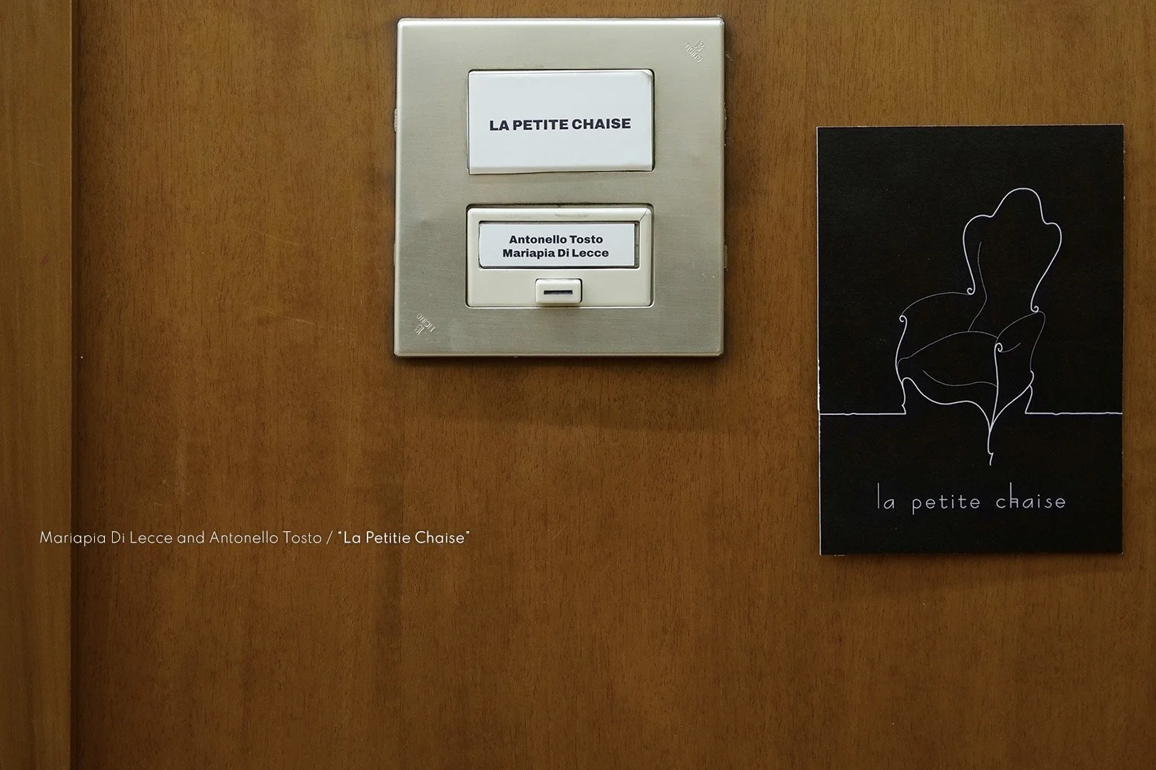 A wall with a silver light switch panel and a black poster. The panel has a white label that says 'LA PETITE CHAISE' and a smaller label with 'Antonello Tosto Mariapia Di Lecce.' The poster features a white outline drawing of a chair and the words 'l