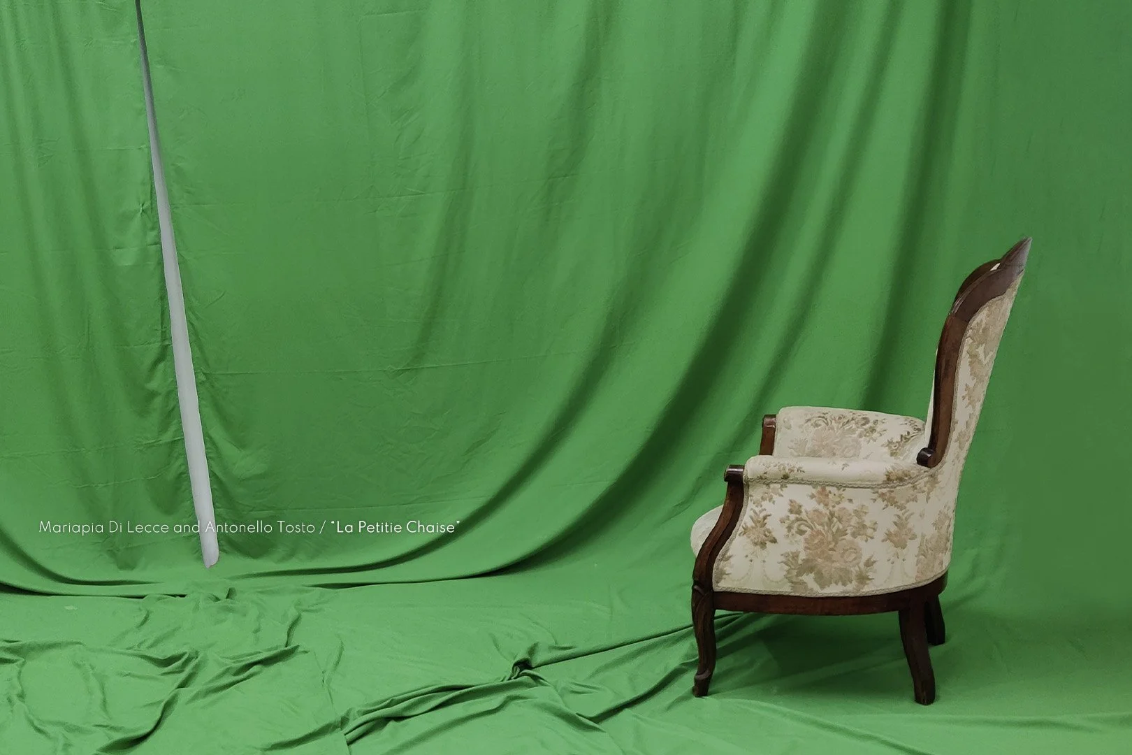 An antique armchair with floral upholstery and wooden legs is positioned to the right on a green fabric backdrop with some overlapping drapery.