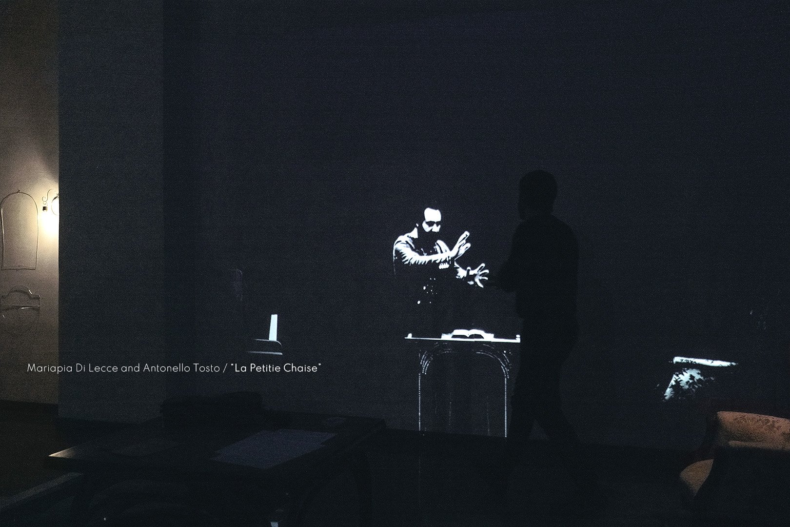 Projection of a man with glasses gesturing with his hands, with a table and a book, in a dark room.