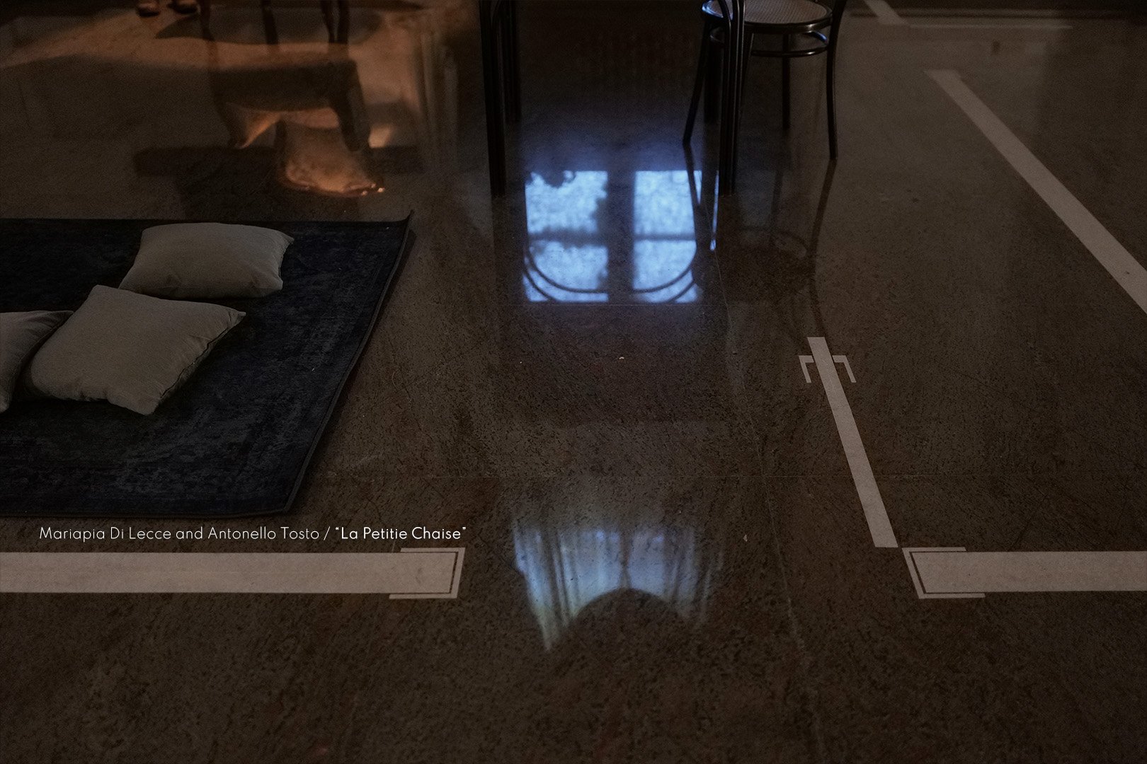 An urban indoor scene with a polished floor reflecting a window with curtains, a small table with chairs, and a dark mat with white pillows on top.