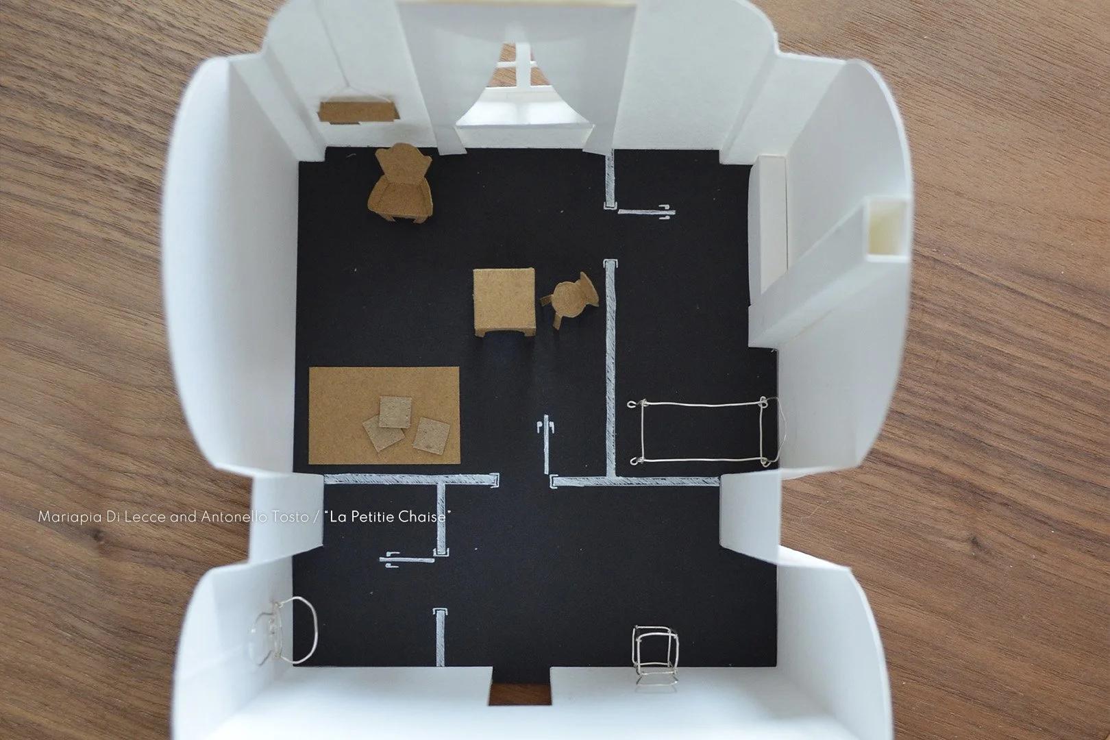 A tabletop architectural model of a small interior space with white walls and black flooring, furnished with miniature furniture including chairs, tables, and a rug.