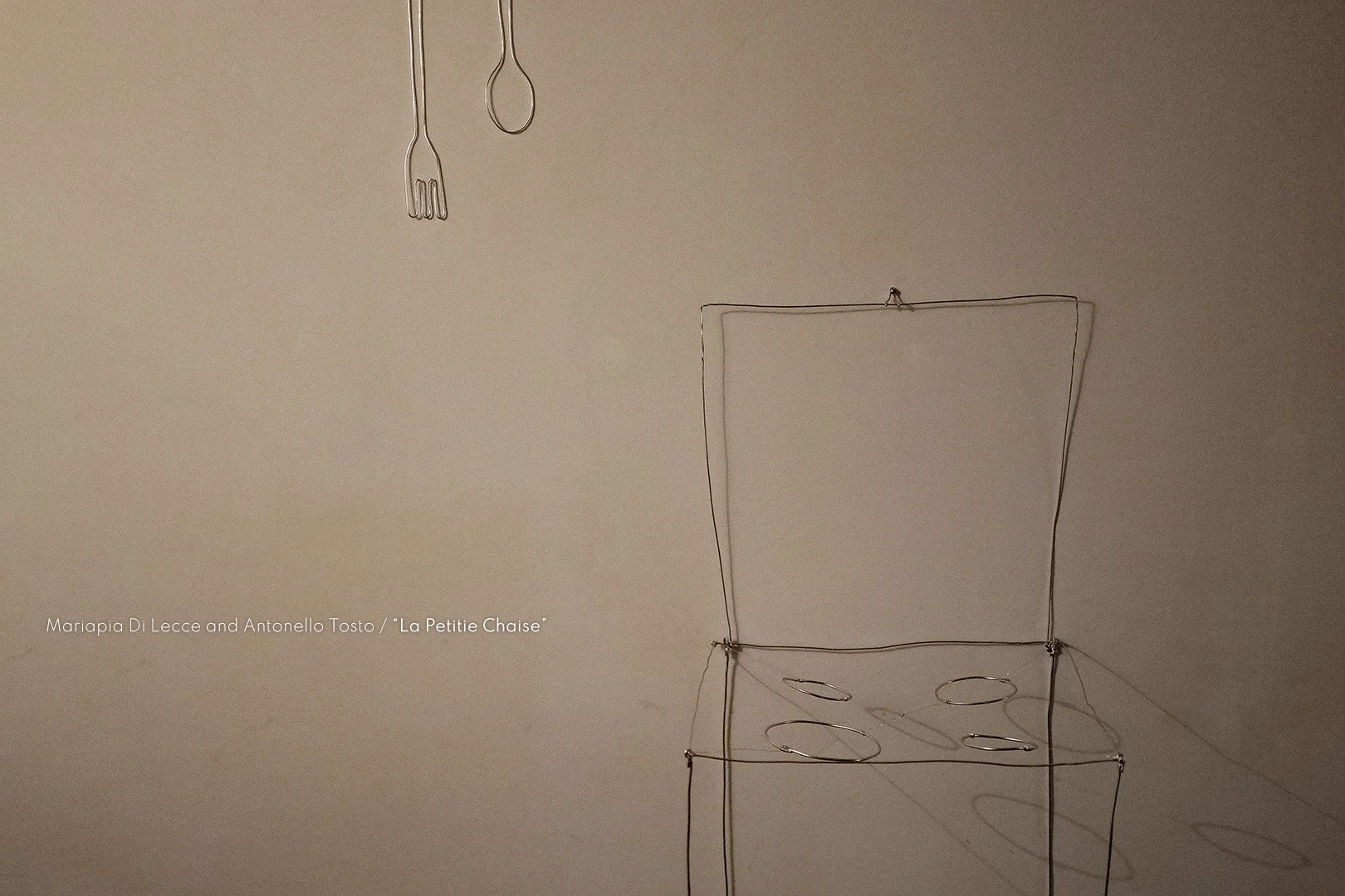 Wire sculpture of a chair with circular cutouts, hanging on a plain wall. Two metal hooks and a fork-shaped wire are also hanging from the ceiling.
