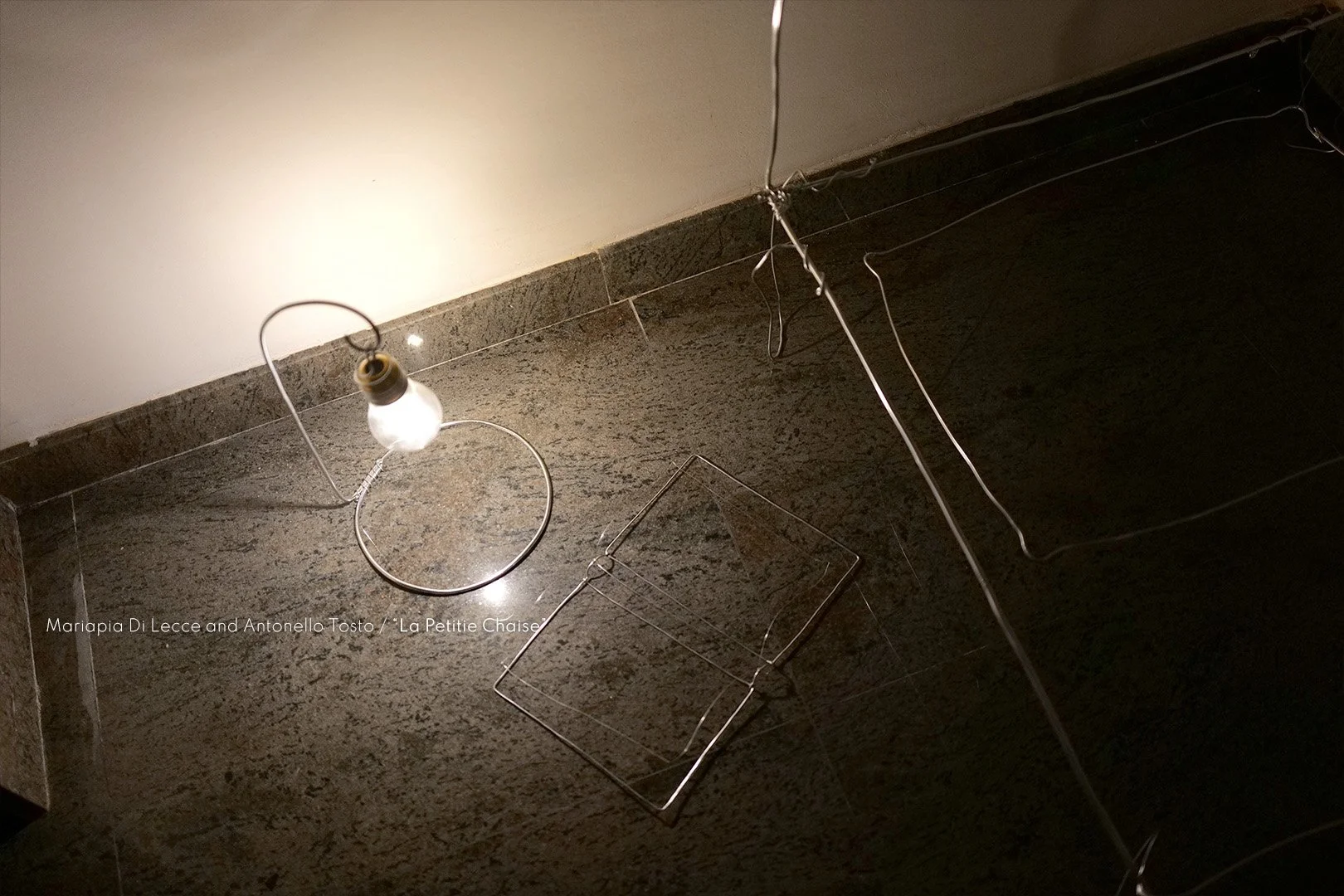 An image of a small light bulb hanging on a wire, next to two wireframe chair models, on a dark granite floor with a wall in the background.