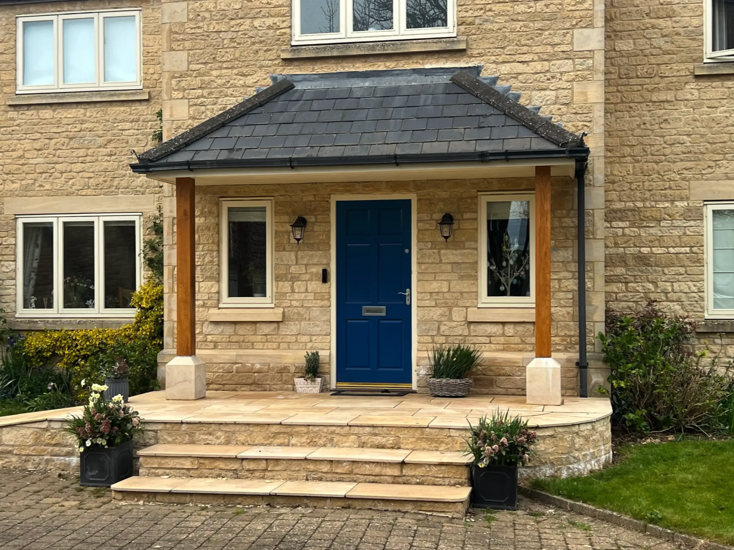 Front porch of a house with a blue door, stone steps, potted plants, and a small covered entryway, with a cobblestone walkway and greenery around.