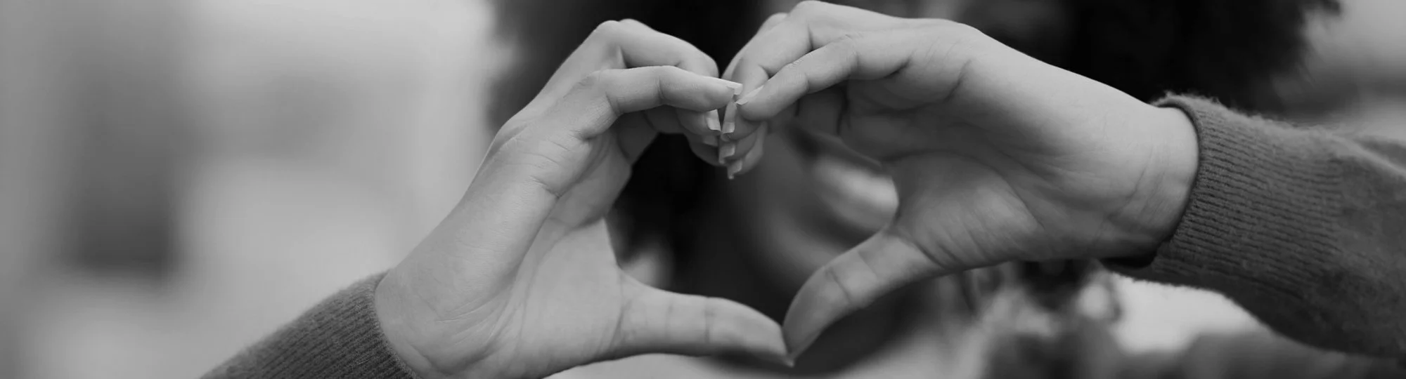 Two hands forming a heart shape with fingers, one hand with a sleeve and the other with a sweater, with a blurred outdoor background.