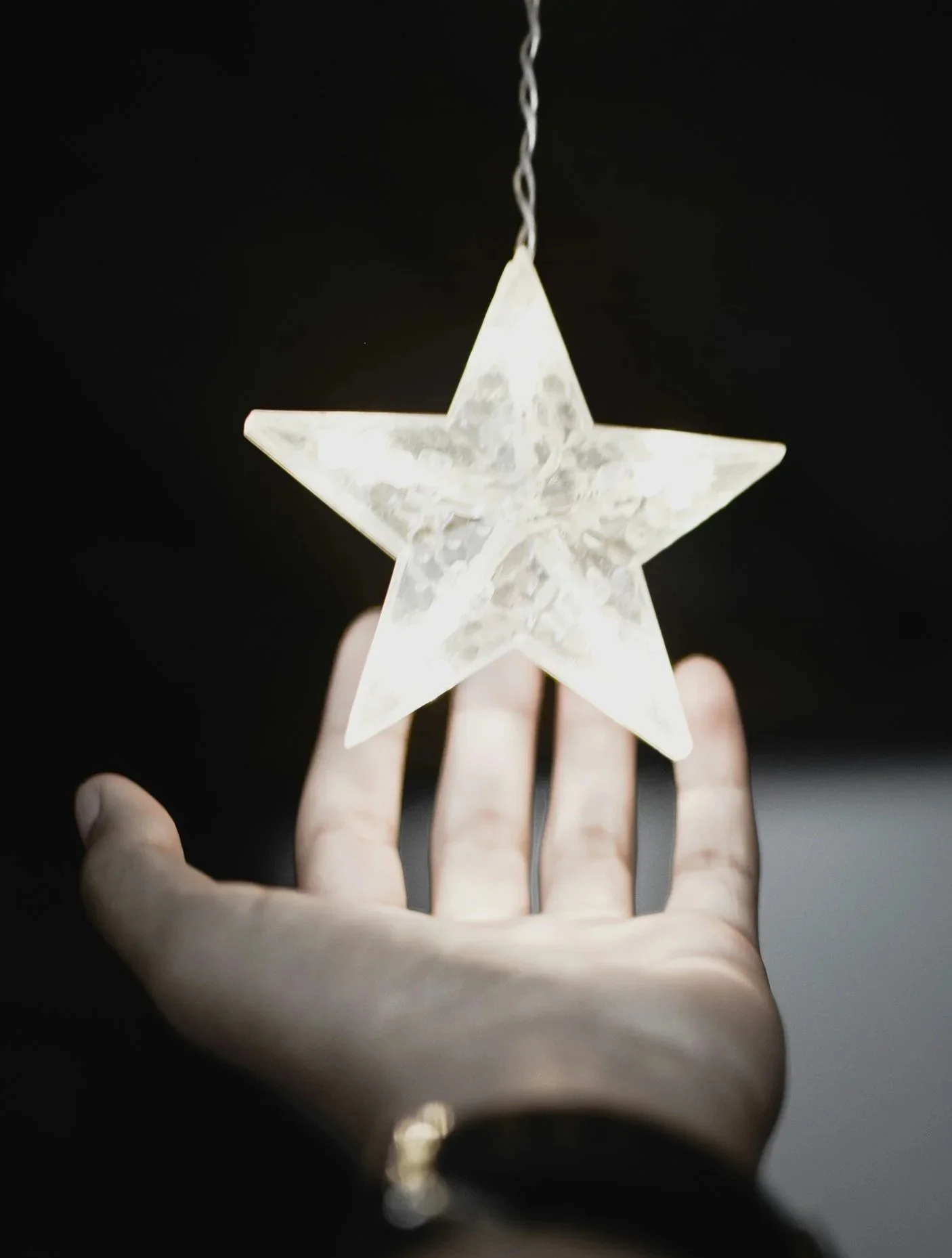 Illuminated star-shaped ornament hanging from a chain, held by a hand with a watch, against a dark background.