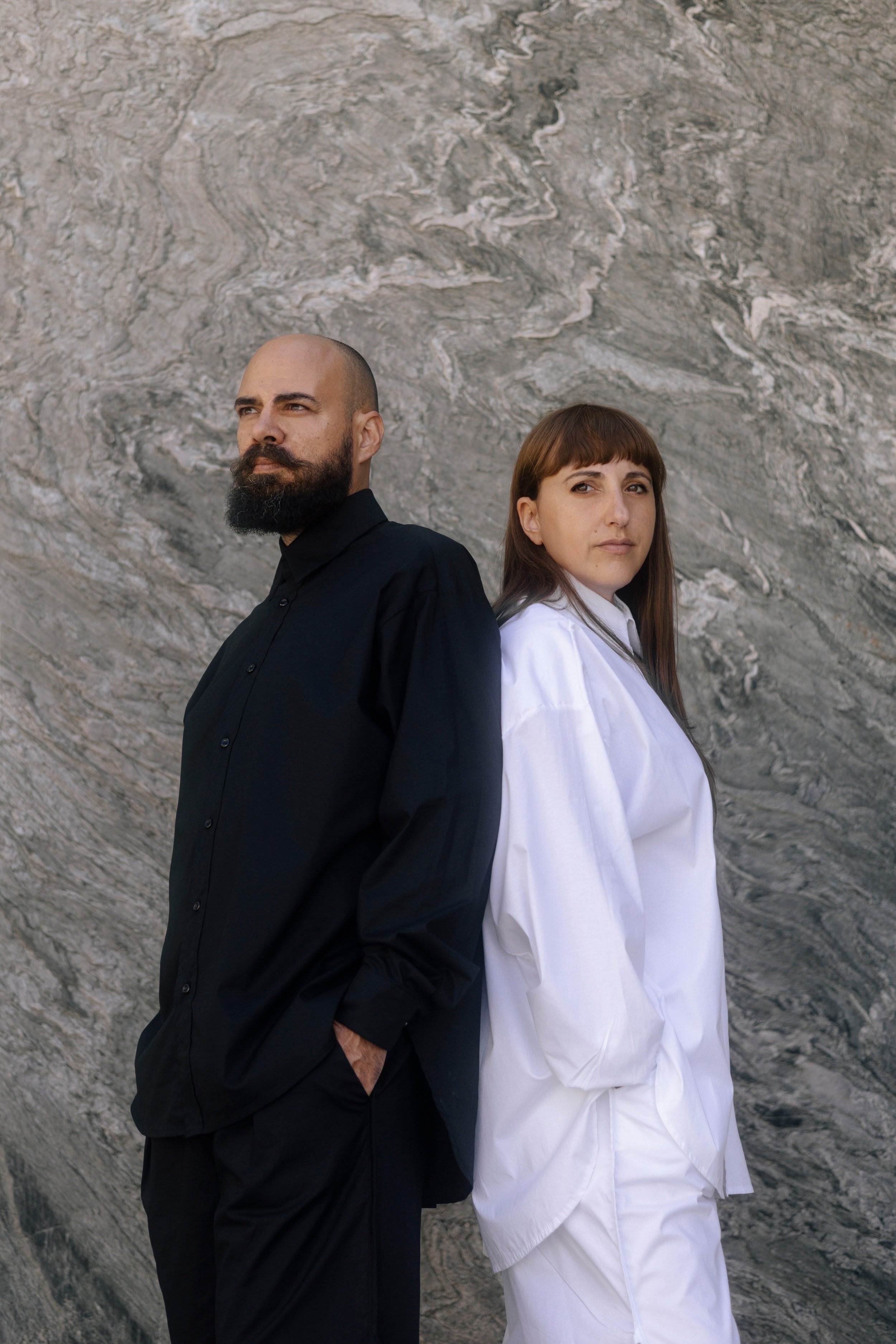 A man with a beard and mustache wearing a black shirt stands back to back with a woman with brown hair wearing a white shirt, both looking away from each other, against a textured gray background.