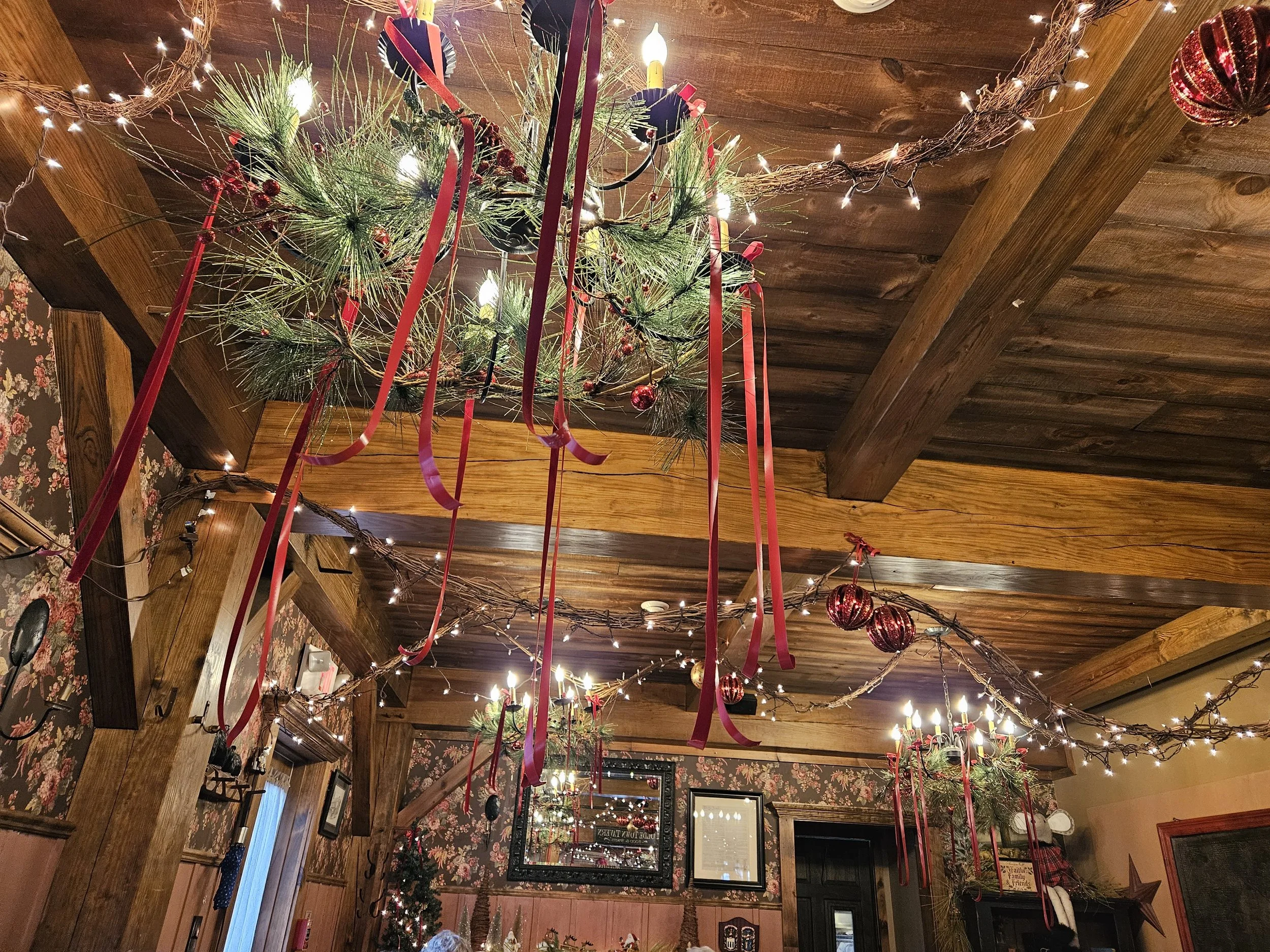 Rustic interior decorated for Christmas with pine garlands, fairy lights, and red ribbons. Ceiling features wood beams and hanging ornaments.