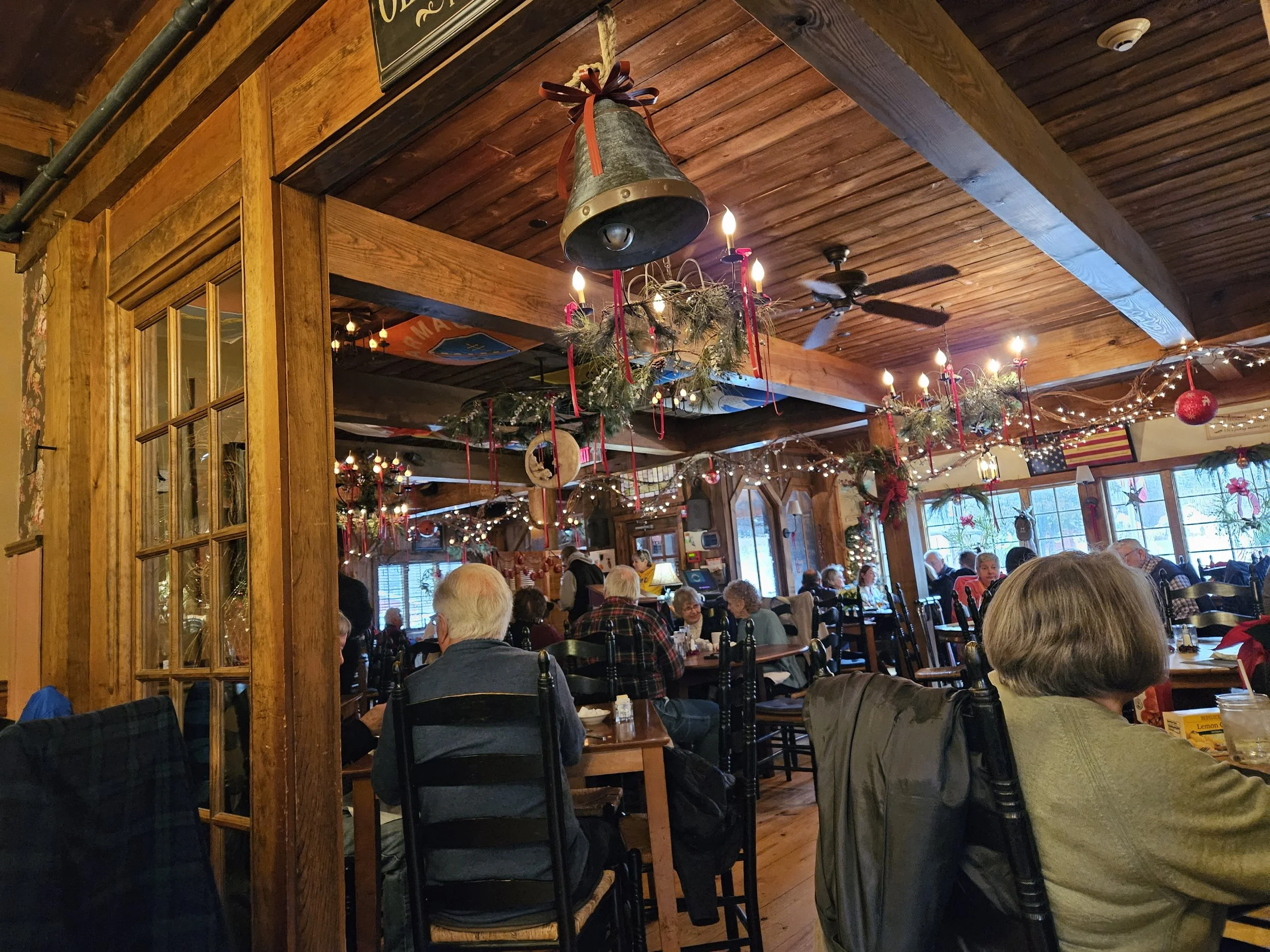 A cozy, rustic-style restaurant interior with wooden decor and holiday decorations, including wreaths and string lights. Patrons are seated at tables, enjoying their meals. A large metal bell hangs from the ceiling, and there are several windows alon