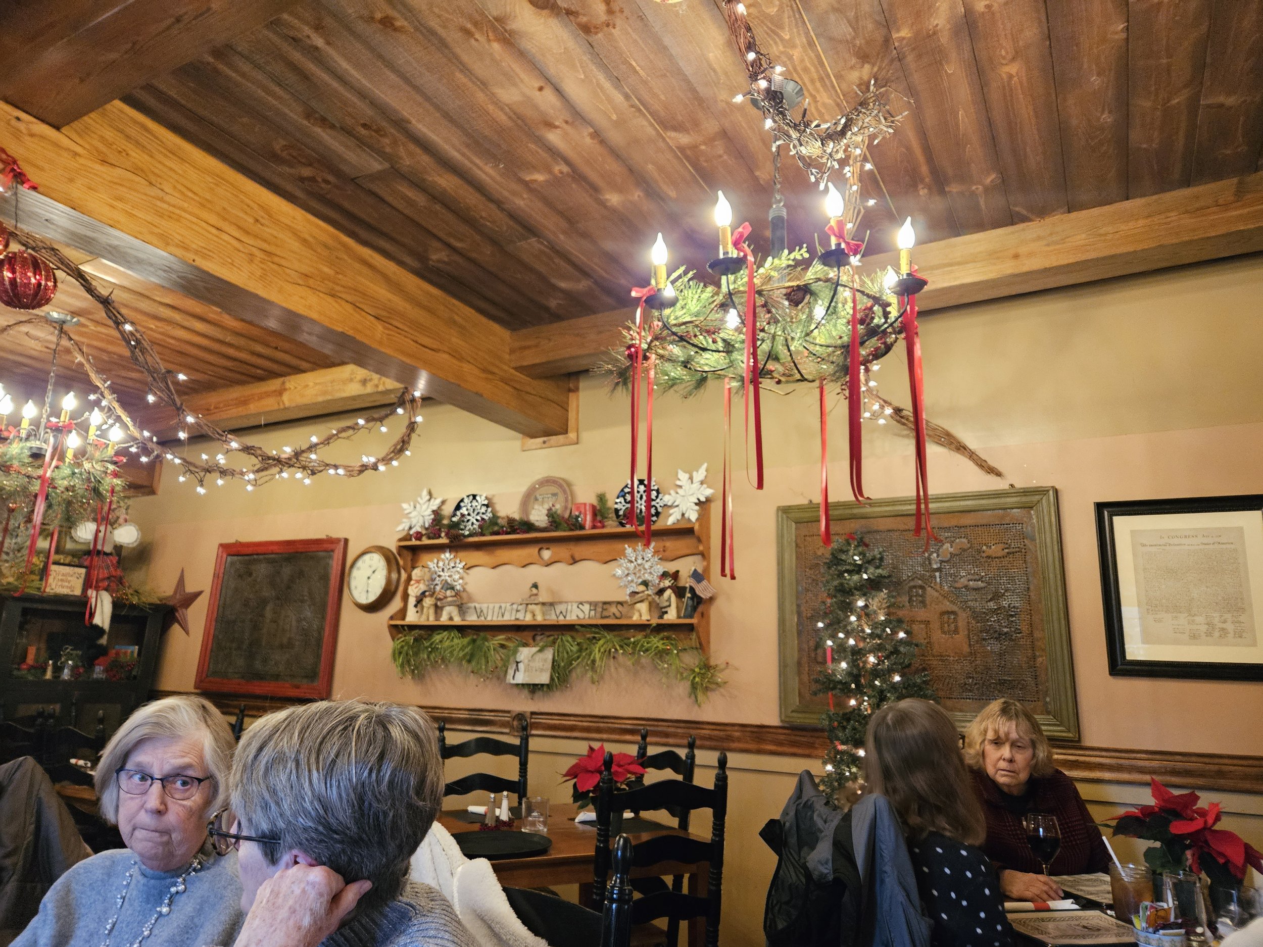 Cozy restaurant interior with holiday decorations, including string lights and red ribbons. Wooden ceiling and walls, framed art, and a small Christmas tree. Two pairs of people seated at tables, engaging in conversation.