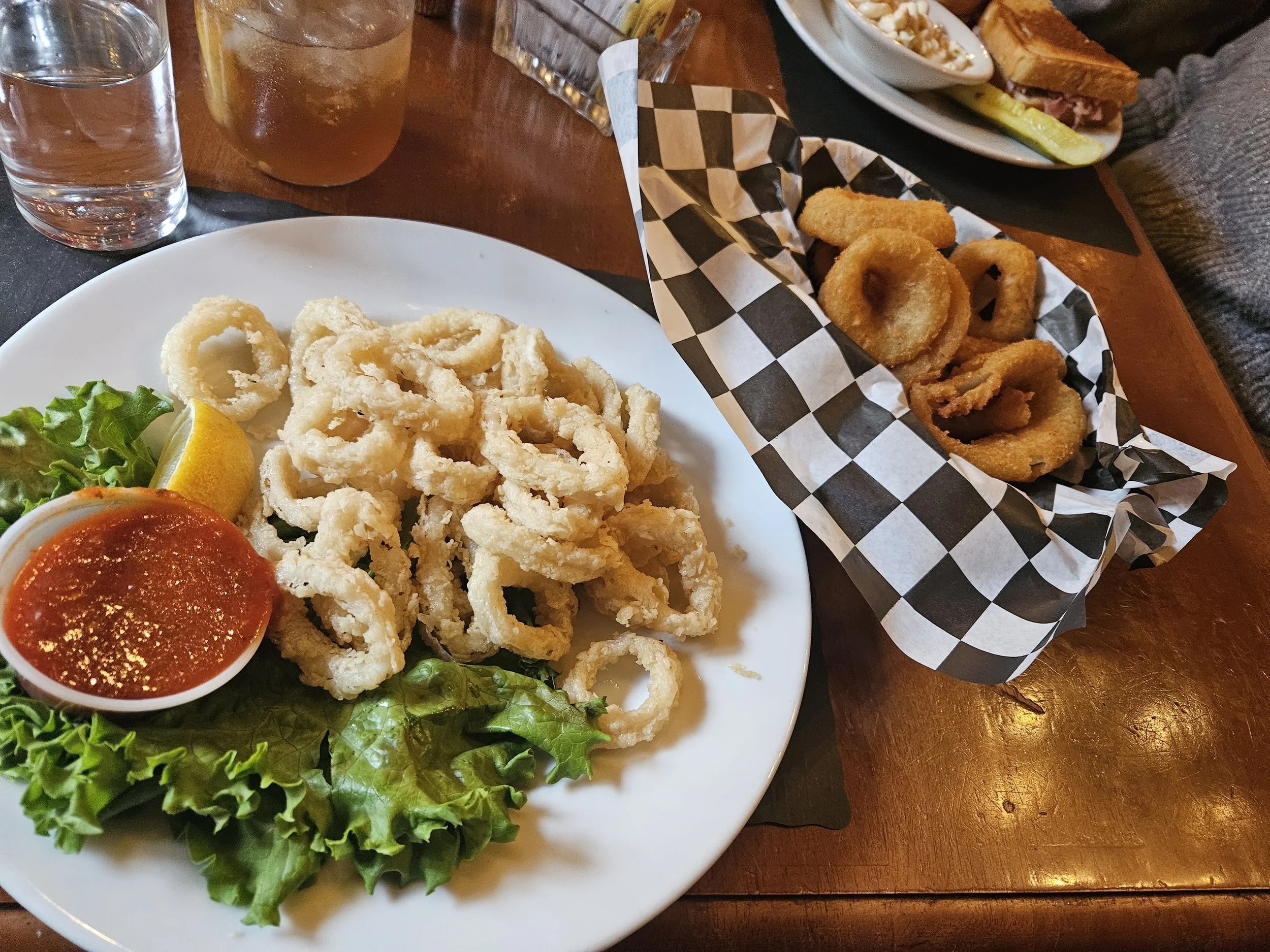 Fried calamari on a plate with marinara sauce and lettuce, alongside a lemon wedge and a separate basket of onion rings on checkered paper. In the background, there's a glass of iced drink and a sandwich with a pickle.