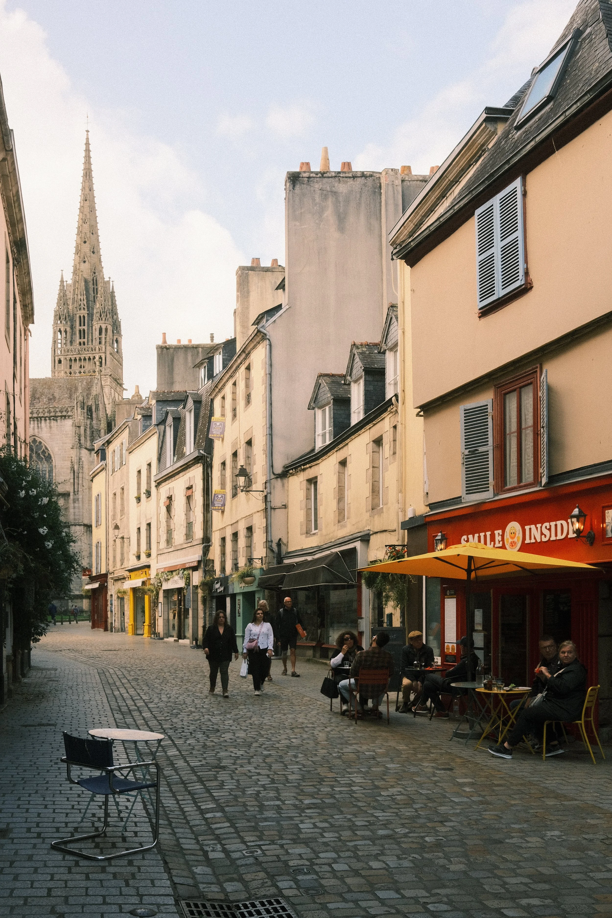 Straße in einer europäischen Stadt mit Cafés und Geschäften, Menschen gehen entlang, im Hintergrund ein gotischer Kirchturm.