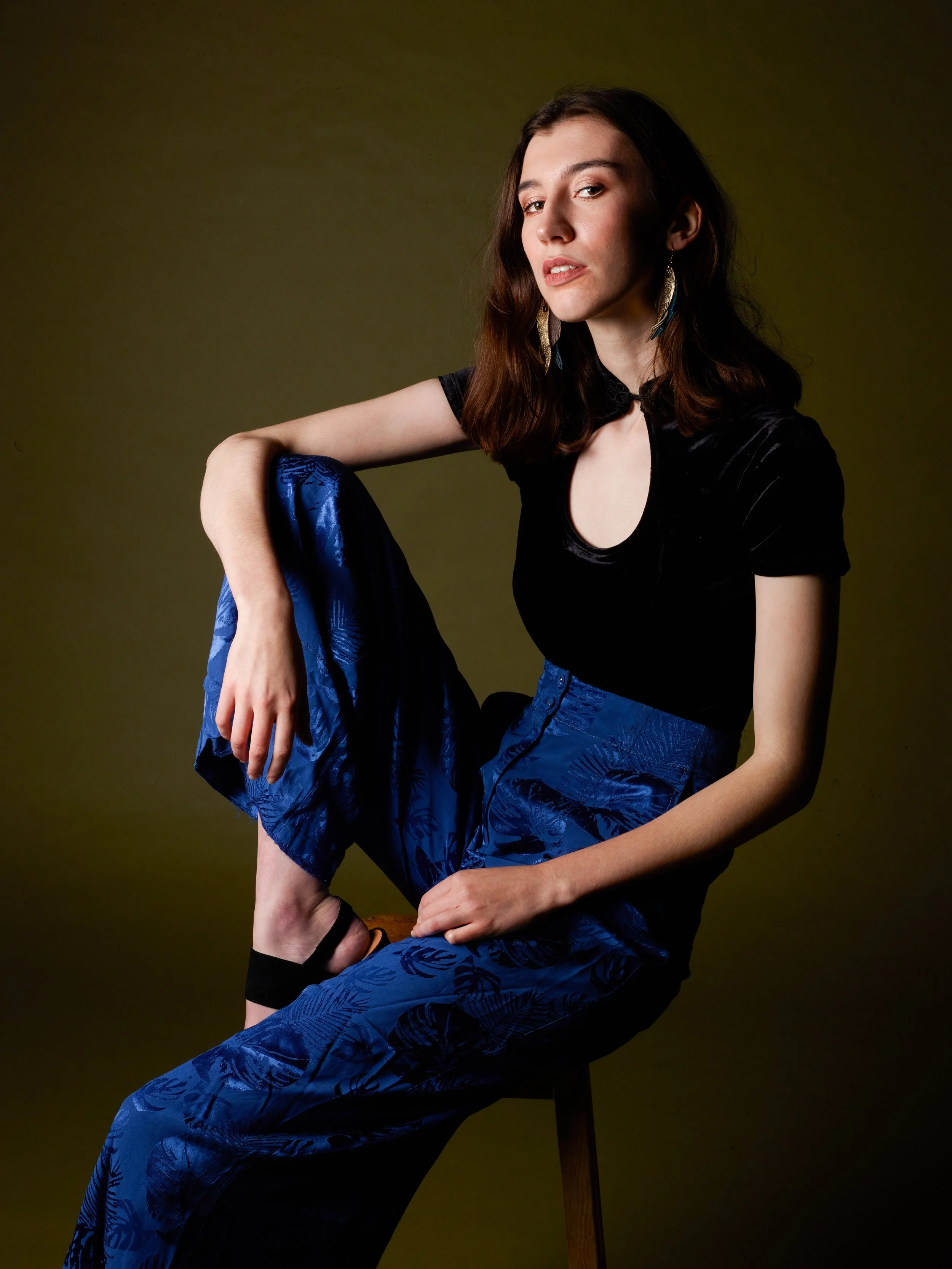 A woman with shoulder-length brown hair, wearing a black top and blue patterned pants, sitting on a stool with a dark background.