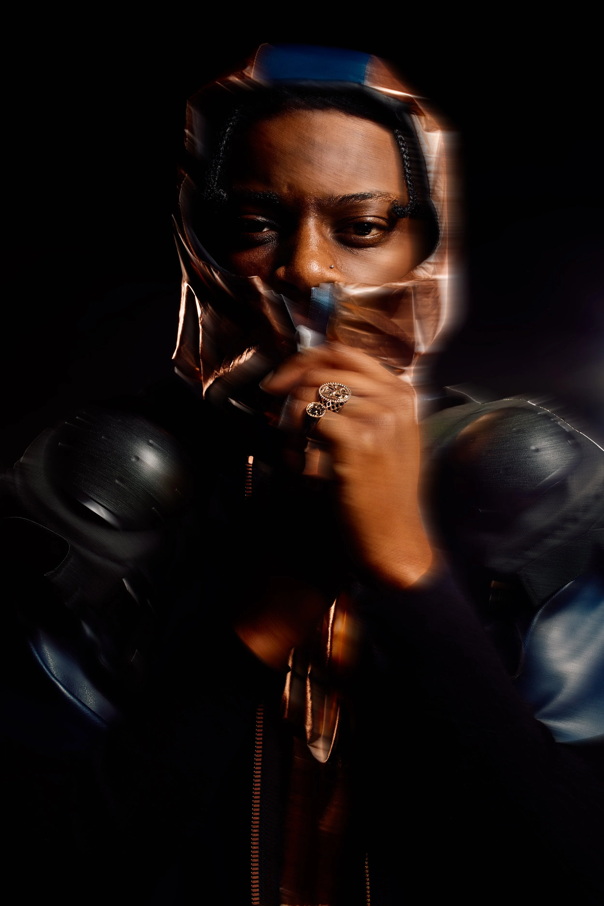 A person with dark skin and braided hair is wearing a shiny, black helmet and black protective gear. They have a nose piercing and rings on their finger, and are holding their hand near their face against a dark background.