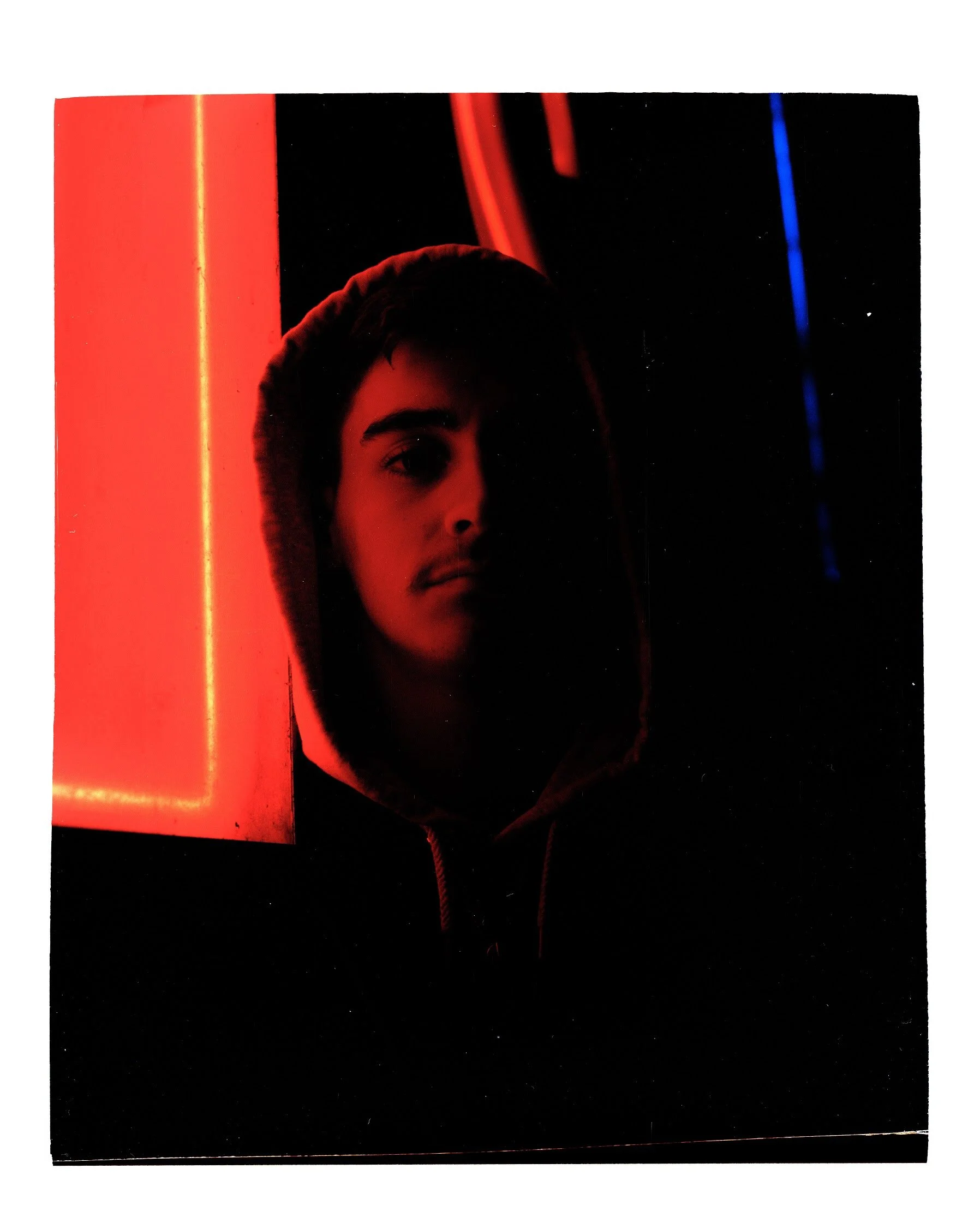 A young man with dark hair, wearing a hoodie, is visible in a dimly lit environment with neon red and blue lights reflected on a nearby surface, creating a cool, urban atmosphere.