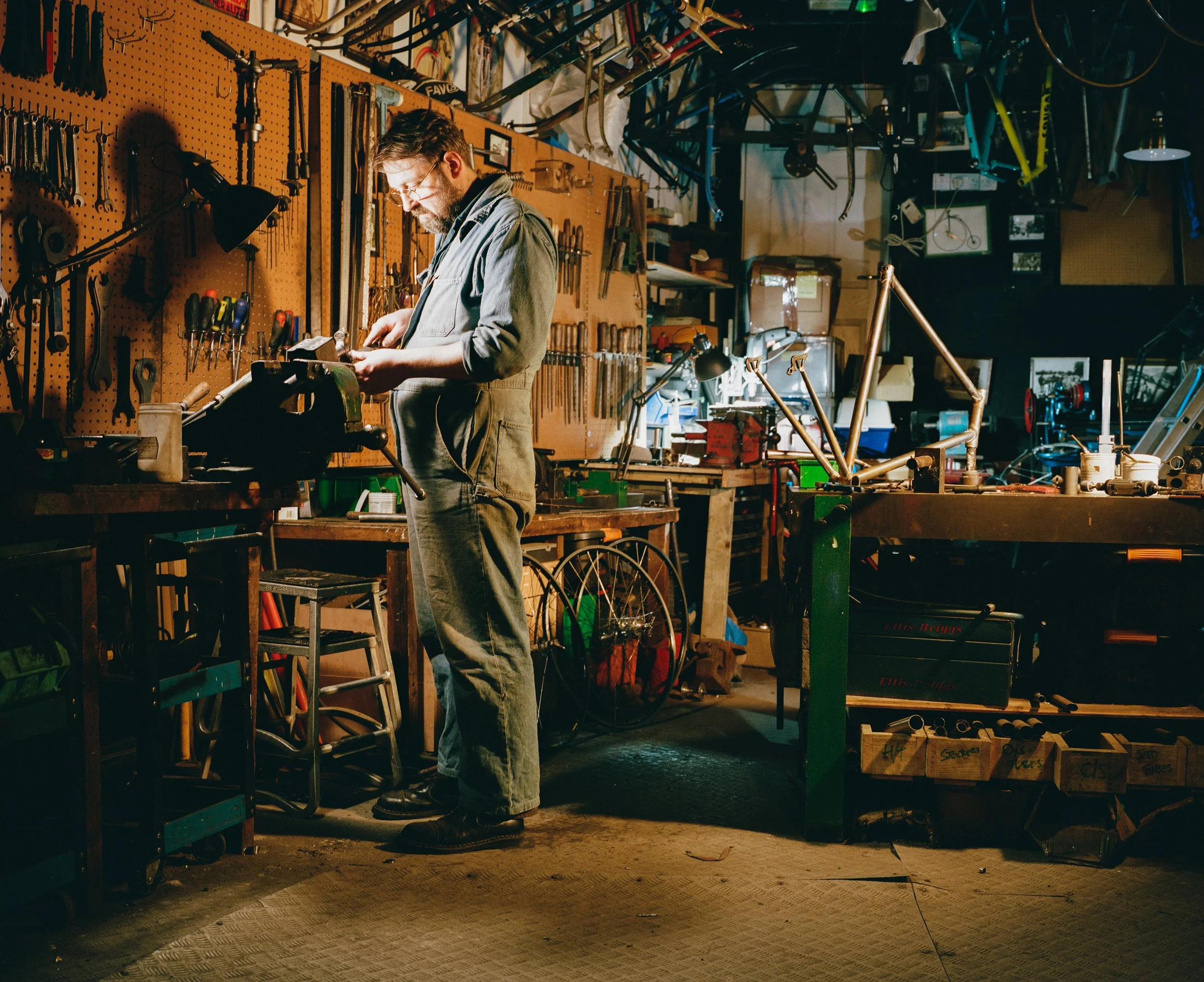 A man in a workshop working on a bicycle frame, surrounded by tools and bicycle parts, with a pegboard wall and tools hanging in the background.