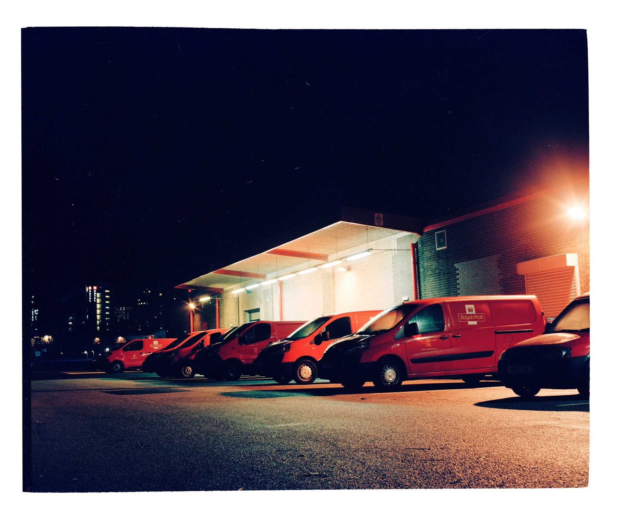 Nighttime scene of a parking lot with several red and black vehicles parked in front of a building with a peaked roof and fluorescent lighting. Bright light near the right edge illuminates part of the building and cars. The dark sky above features visible stars and faint streaks of light, possibly from passing aircraft or satellites.