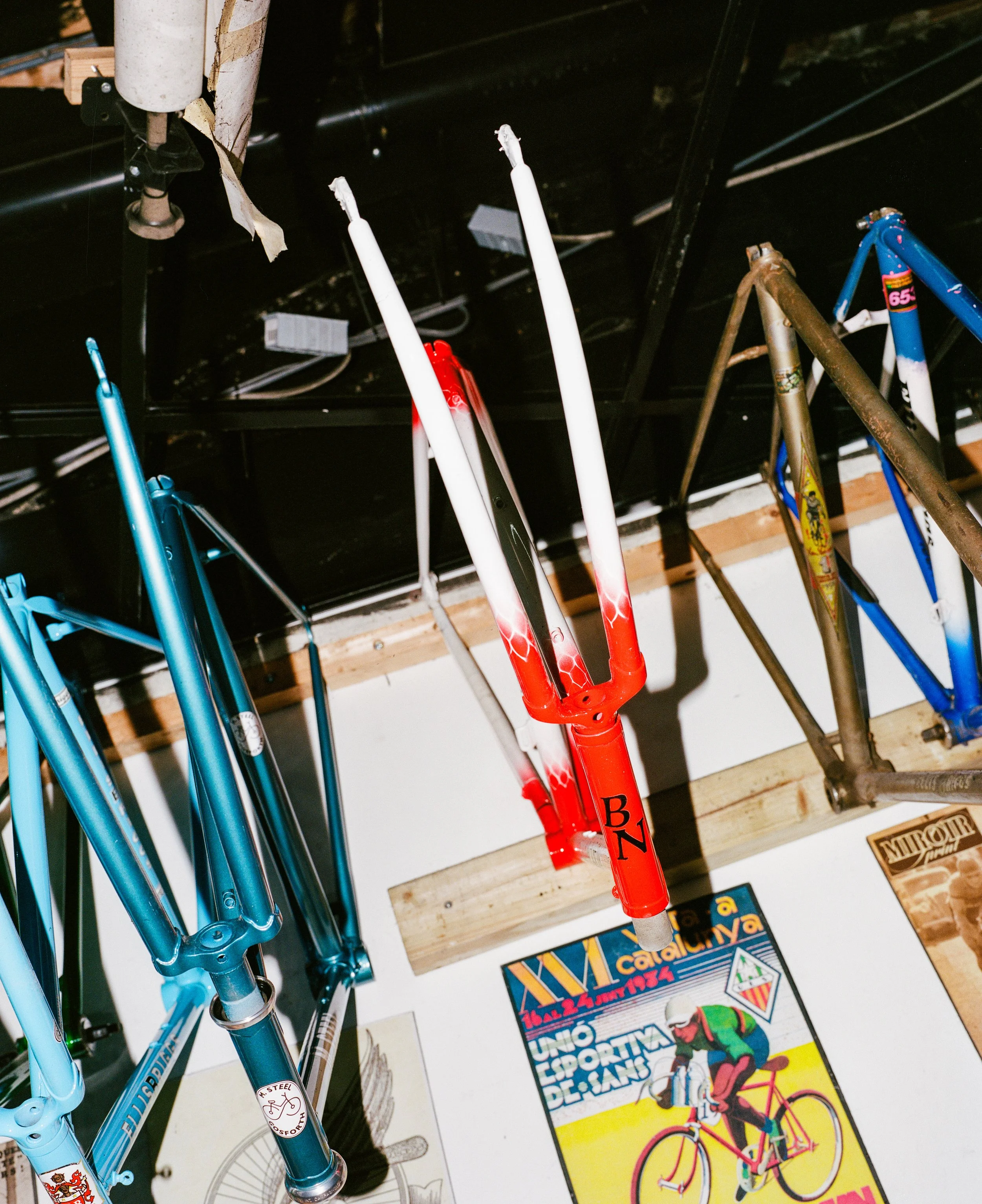 Empty bicycle frames hung on a wall, some painted in bright colors and others rusted, positioned above vintage posters of cycling events.