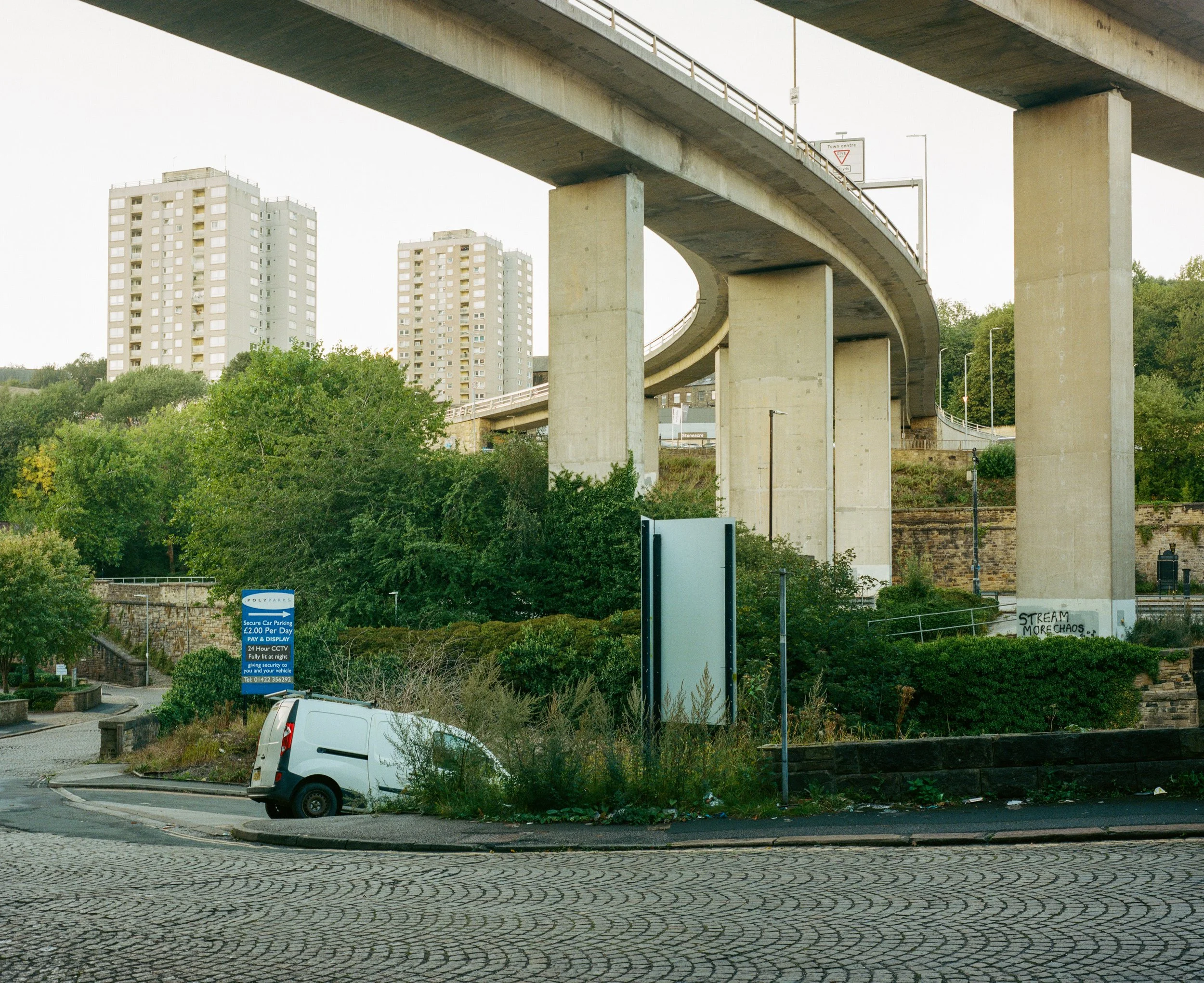Underpass with tall concrete support columns, green trees, multi-story apartment buildings in the background, a car parked at the bottom, and signage including a blue parking notice and a street sign.